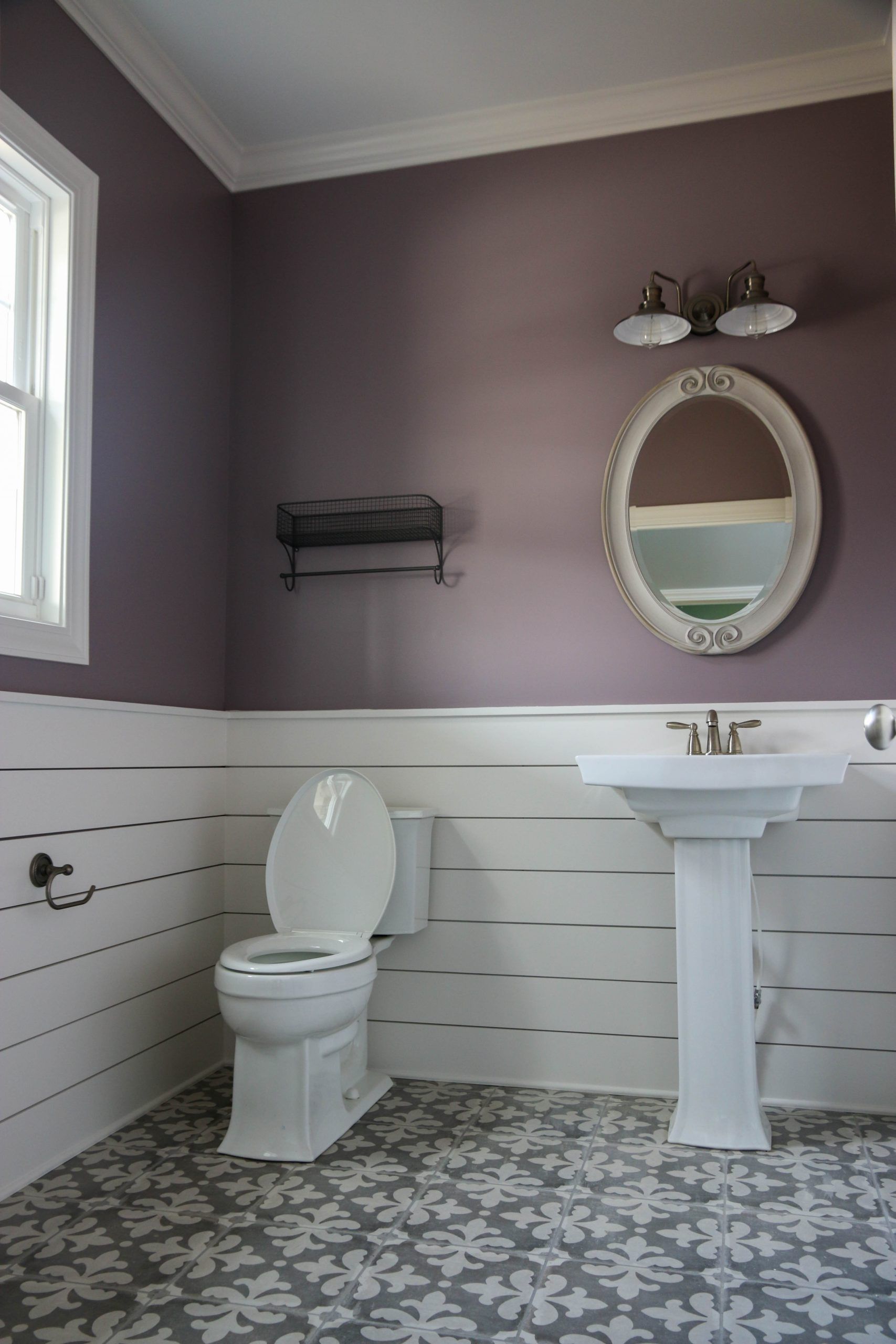 Purple-and-white bathroom with pedestal sink, toilet, oval mirror, and patterned floor tile.