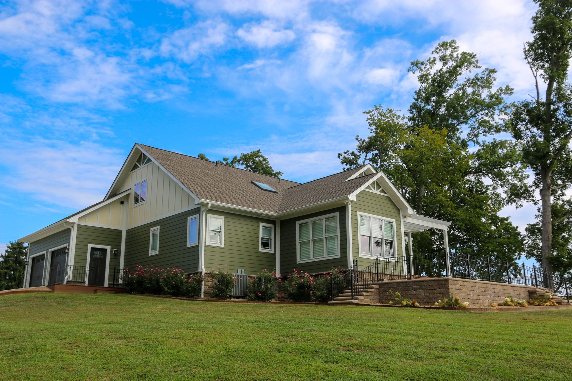 Green house with white trim on a grassy hill under a blue sky, surrounded by trees