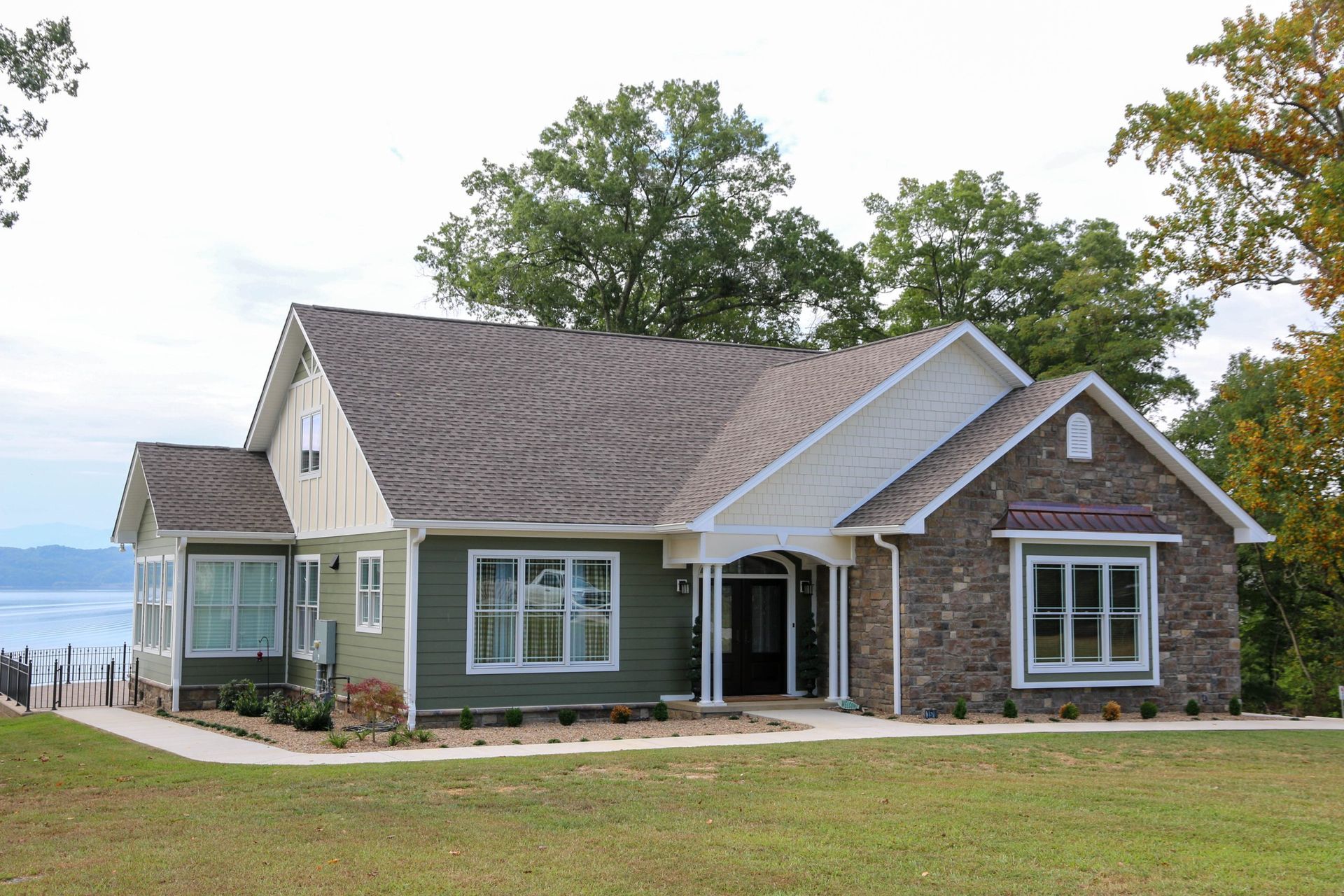 Modern house with gray siding and stone facade overlooking a lake, with a lawn and trees in front