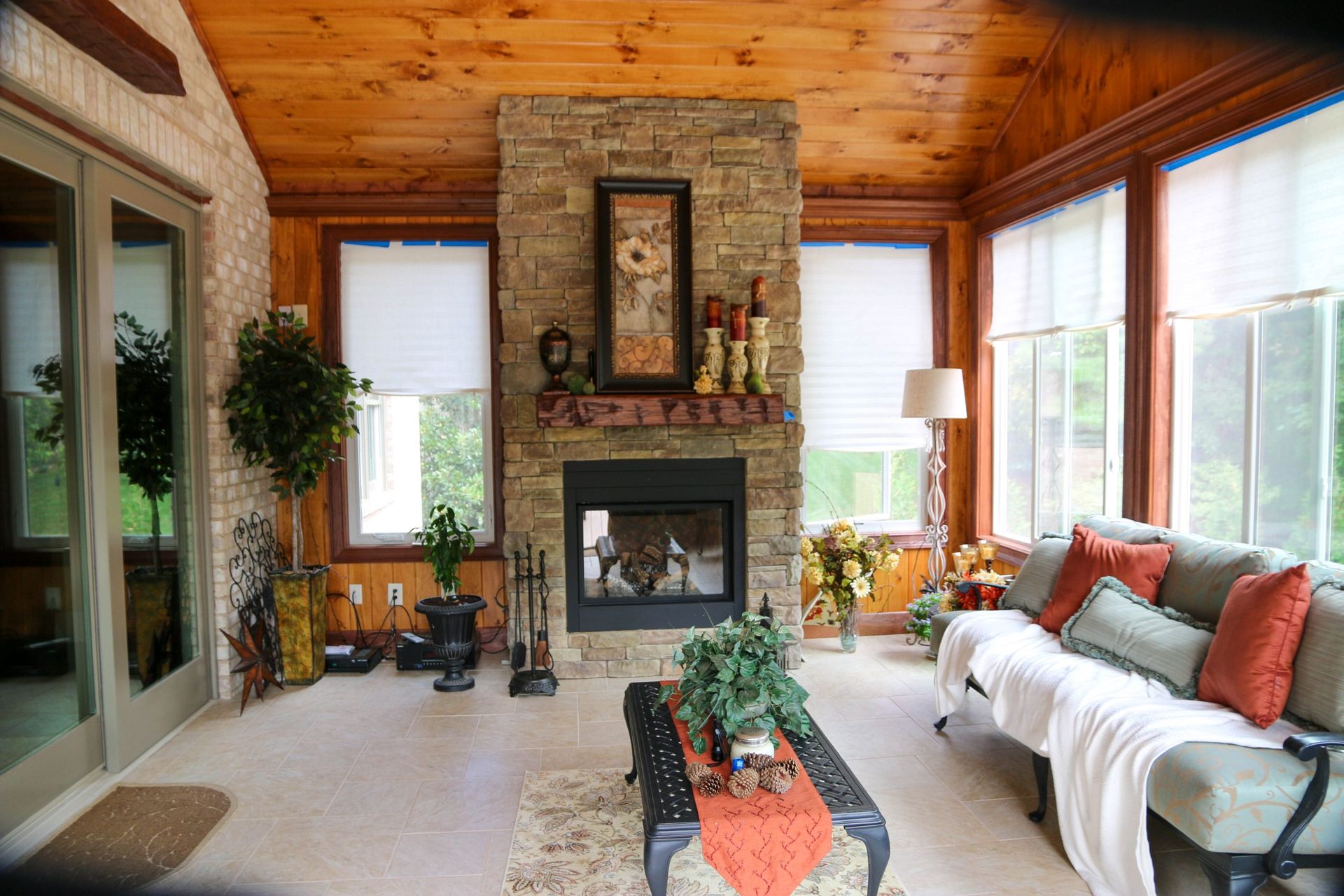 Cozy living room with stone fireplace, wood ceiling, large windows, and white sofa with orange pillows