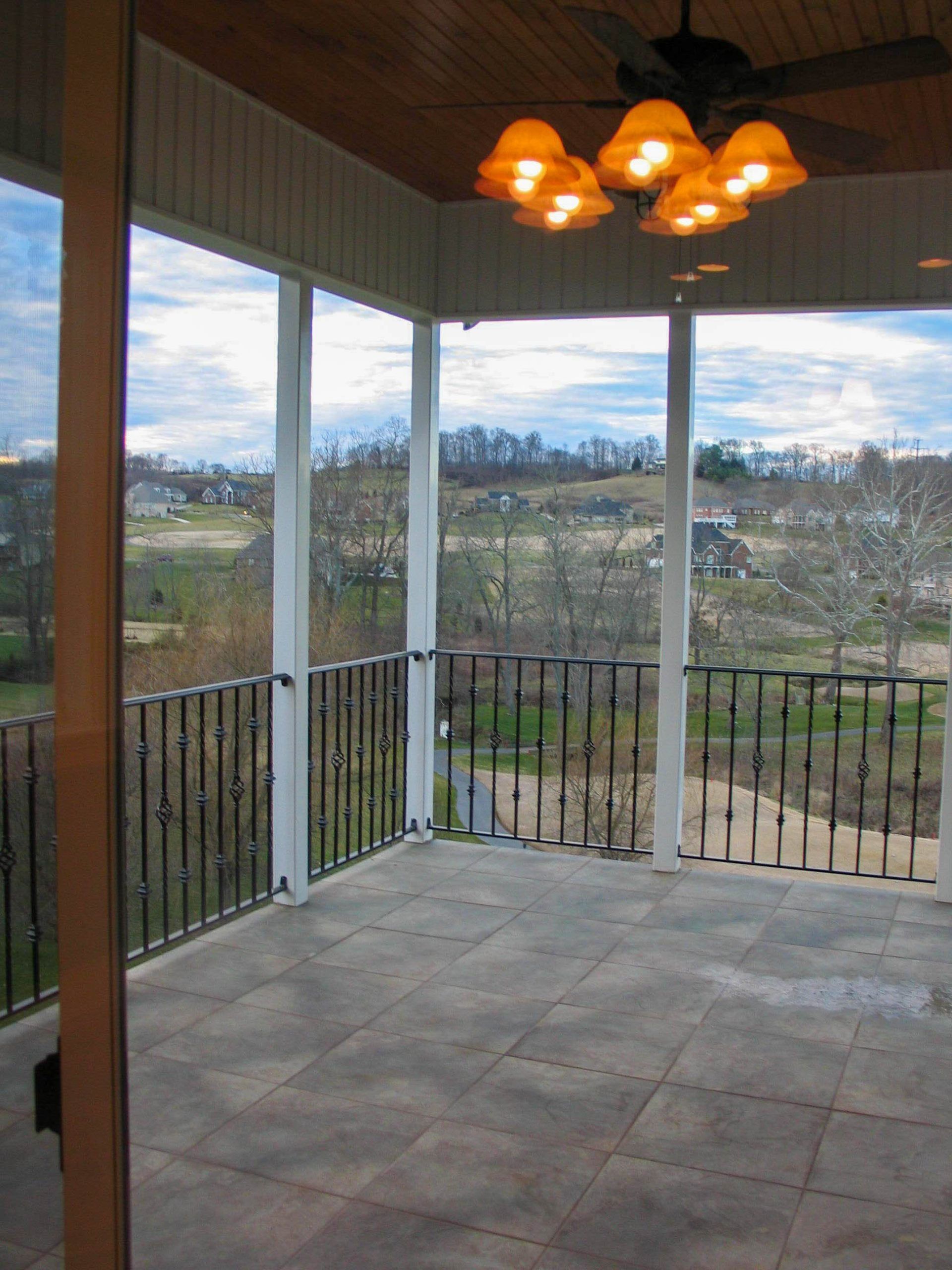 Covered porch with stone floor, black railing, hanging light fixture, and a view of fields and cloudy sky.