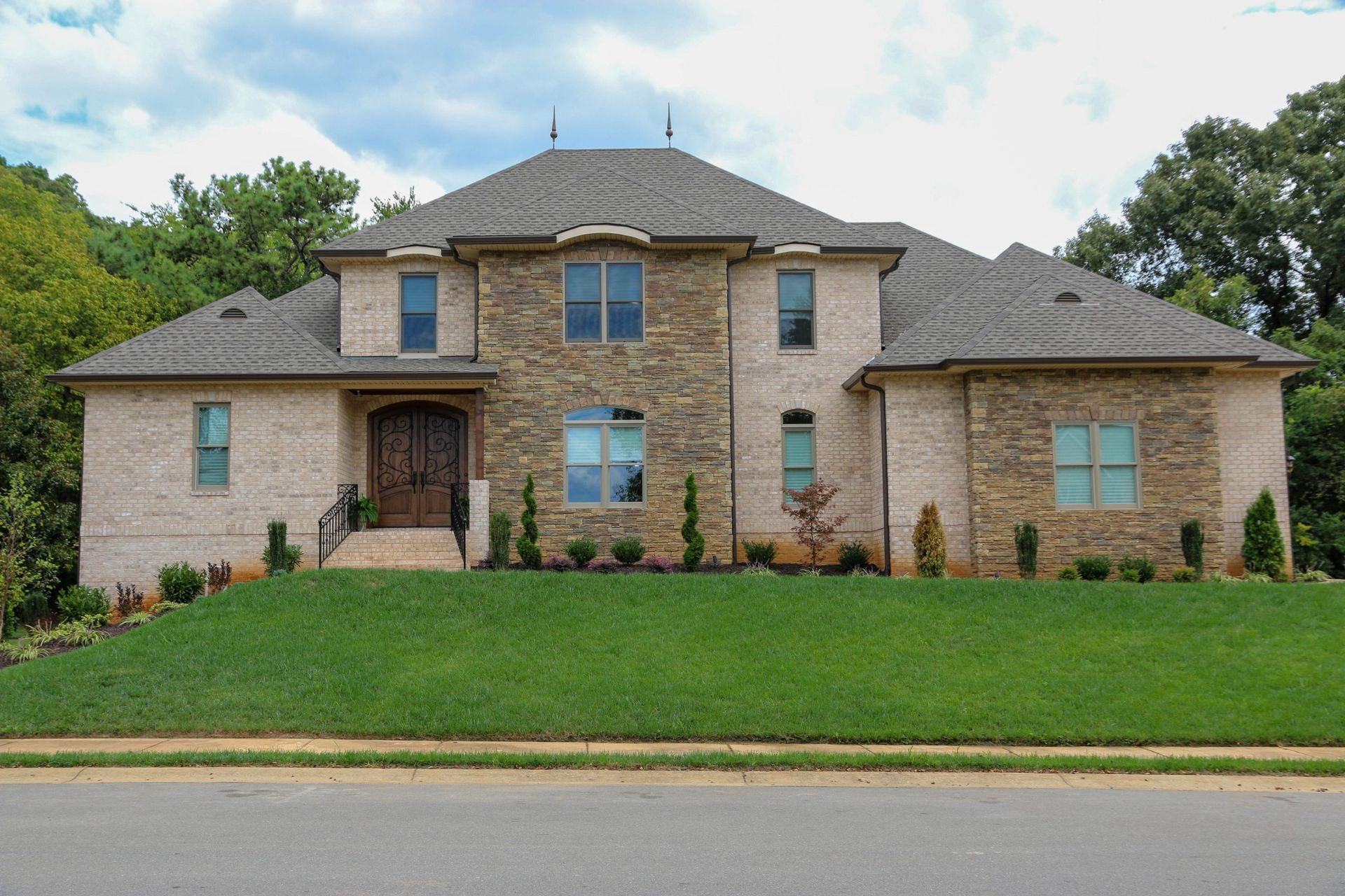 Large two-story stone house with a front lawn, arched entryway, and blue windows under a cloudy sky