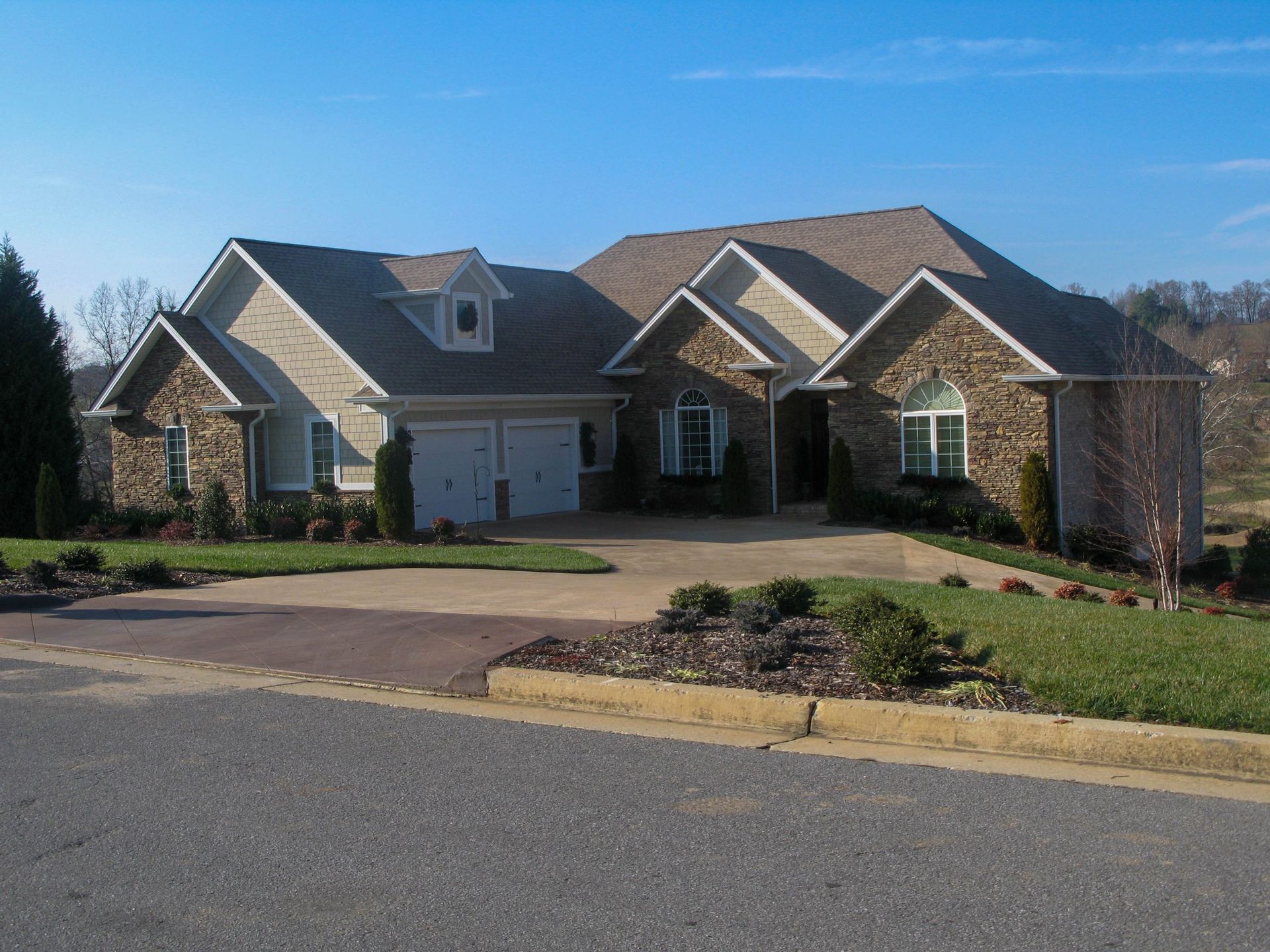 Suburban brick house with three-car garage, landscaped front yard, and curved driveway under a clear blue sky