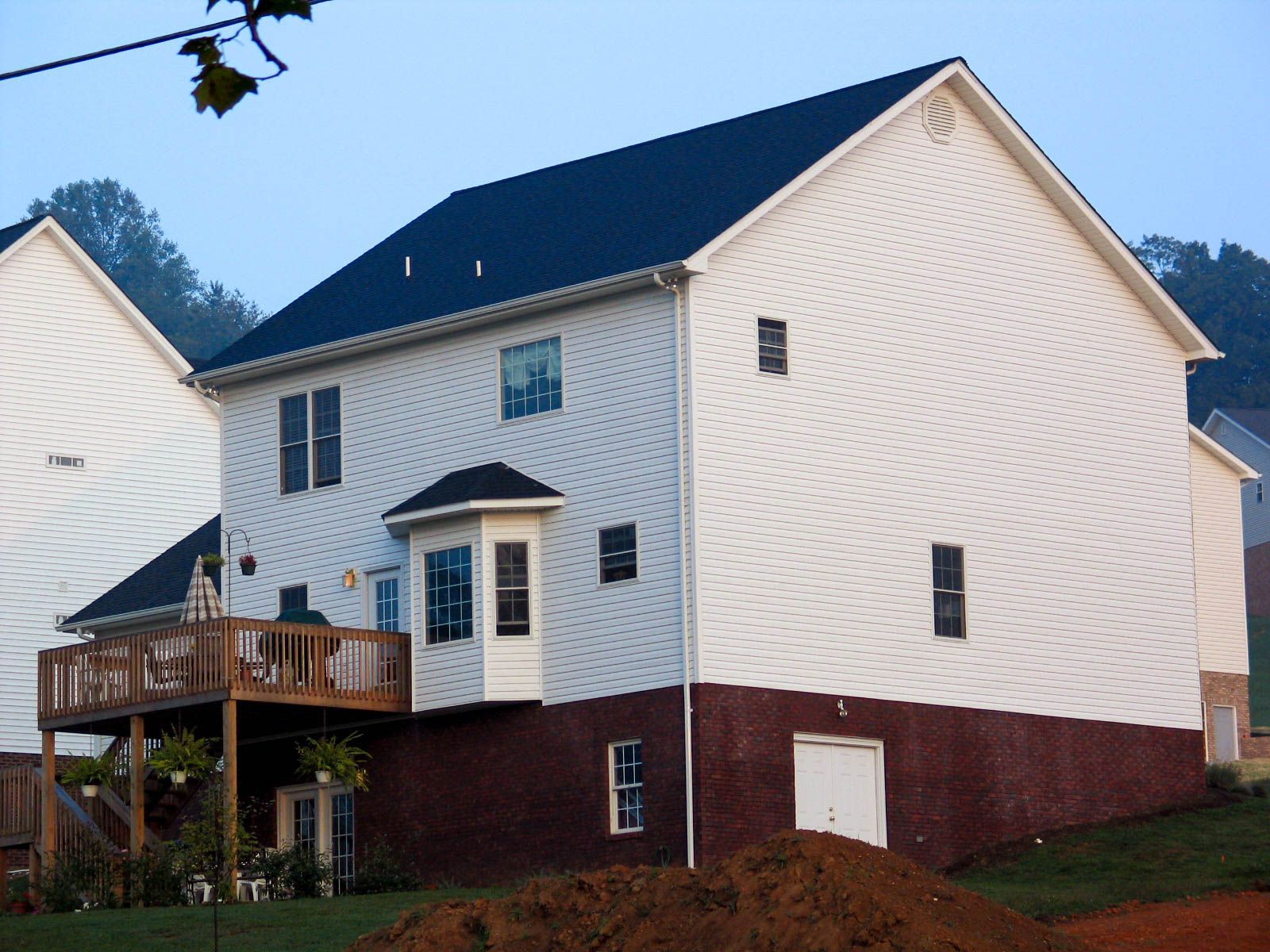 Two-story white house with a dark roof, brick foundation, and wooden deck on a grassy slope.