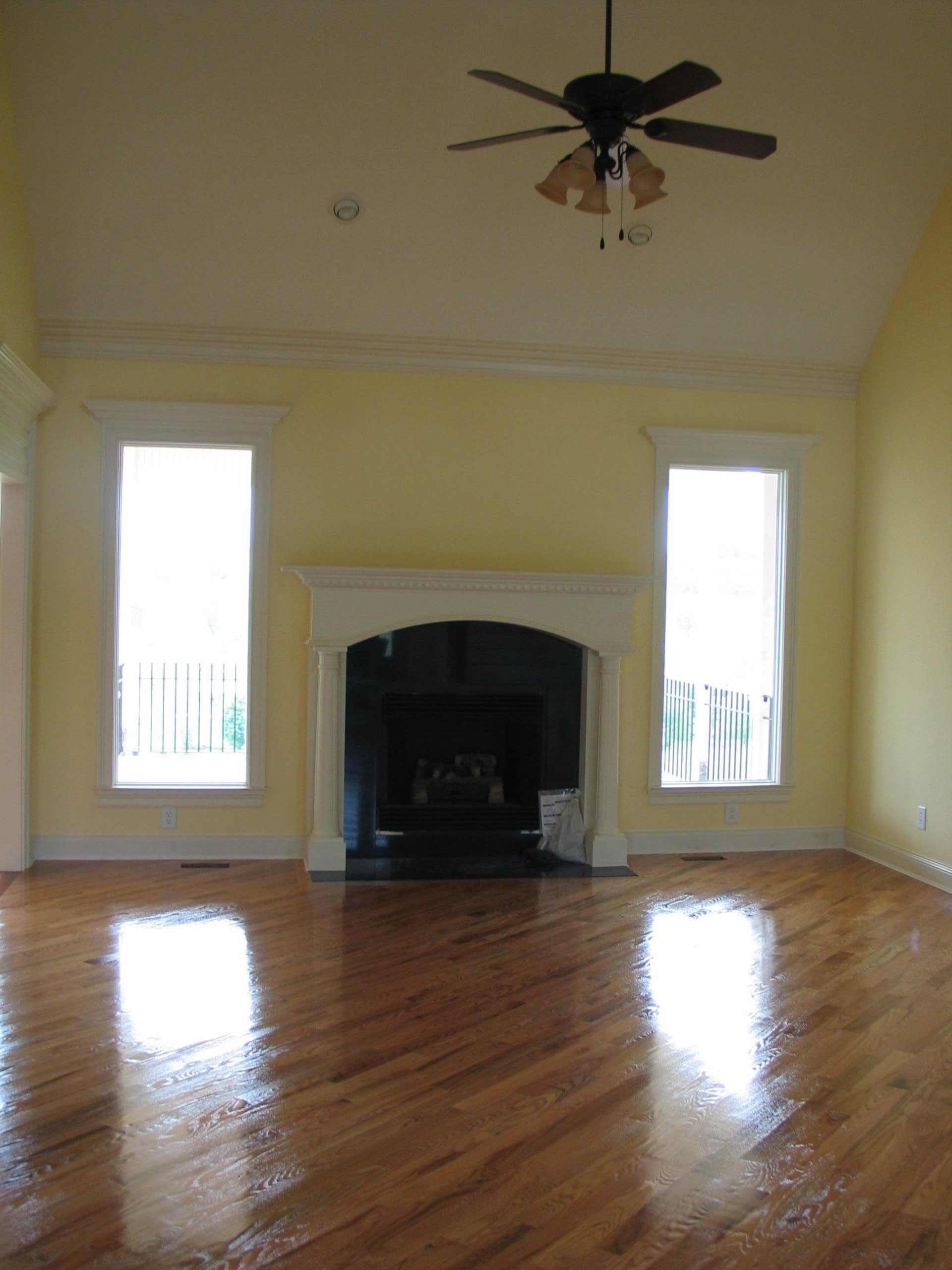 Empty living room with hardwood floors, two tall windows, and a dark fireplace beneath a ceiling fan
