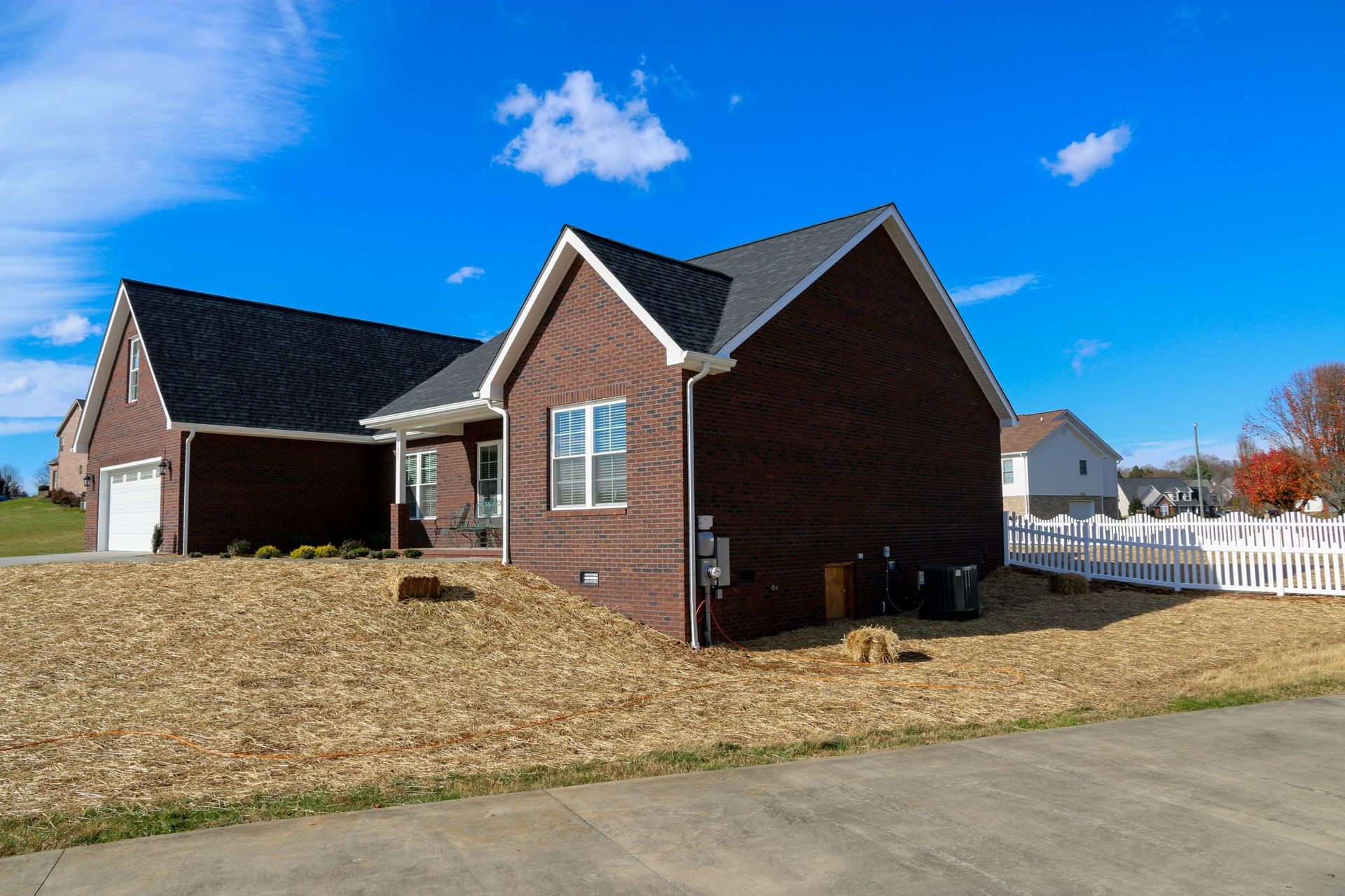 Brick suburban house with attached garage, dark roof, white fence, and blue sky on a grassy lot
