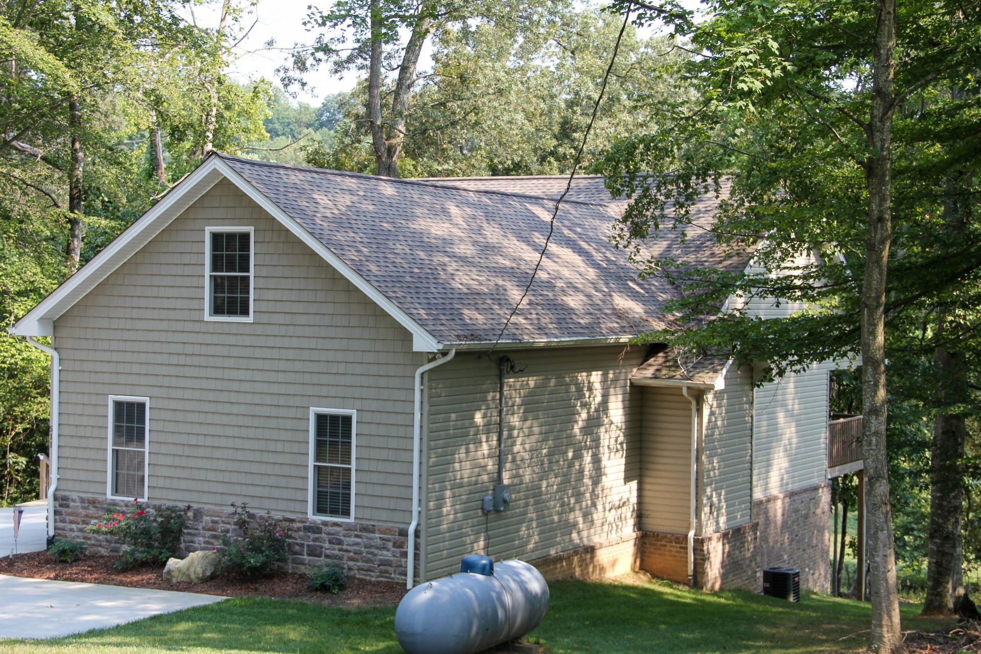 Gray house with tan siding and a metal propane tank in a wooded yard