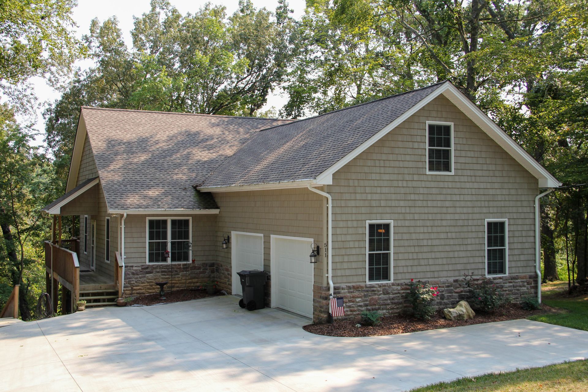 A tan, multi-story house with a stone base and attached two-car garage surrounded by trees on a sunny day.