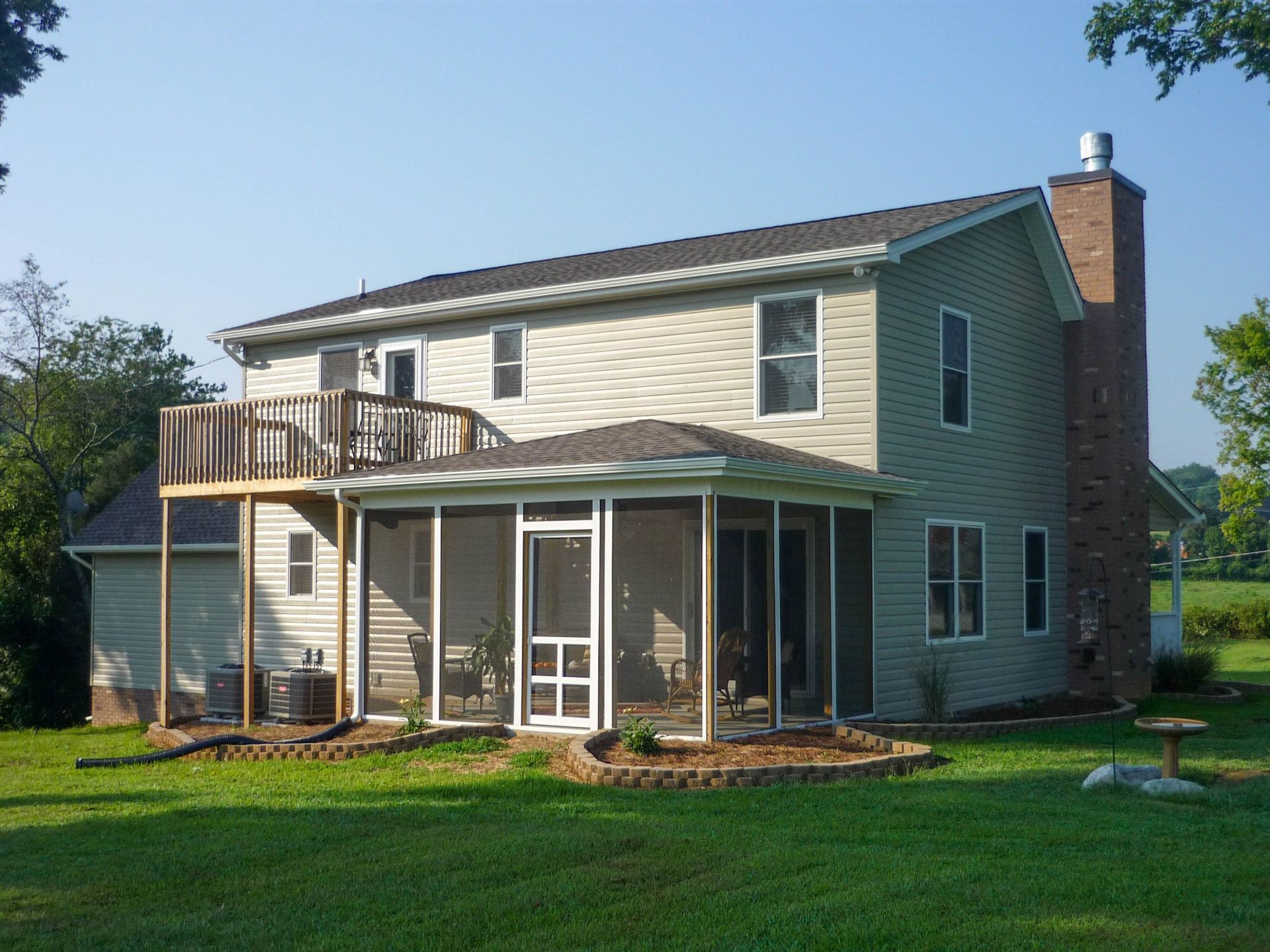 Two-story beige house with screened porch, deck, and green lawn under a clear blue sky