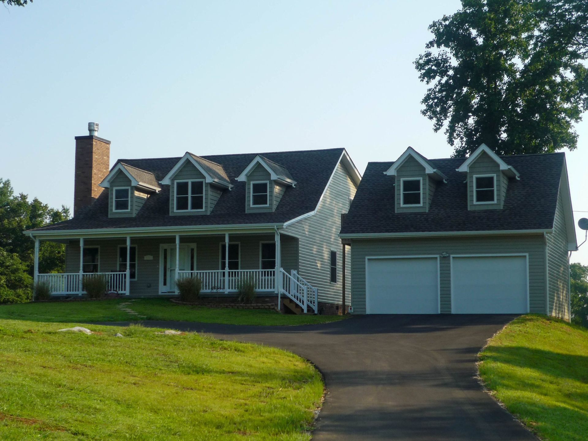 Two-story gray house with dormer windows, front porch, detached two-car garage, and curved driveway in a grassy yard