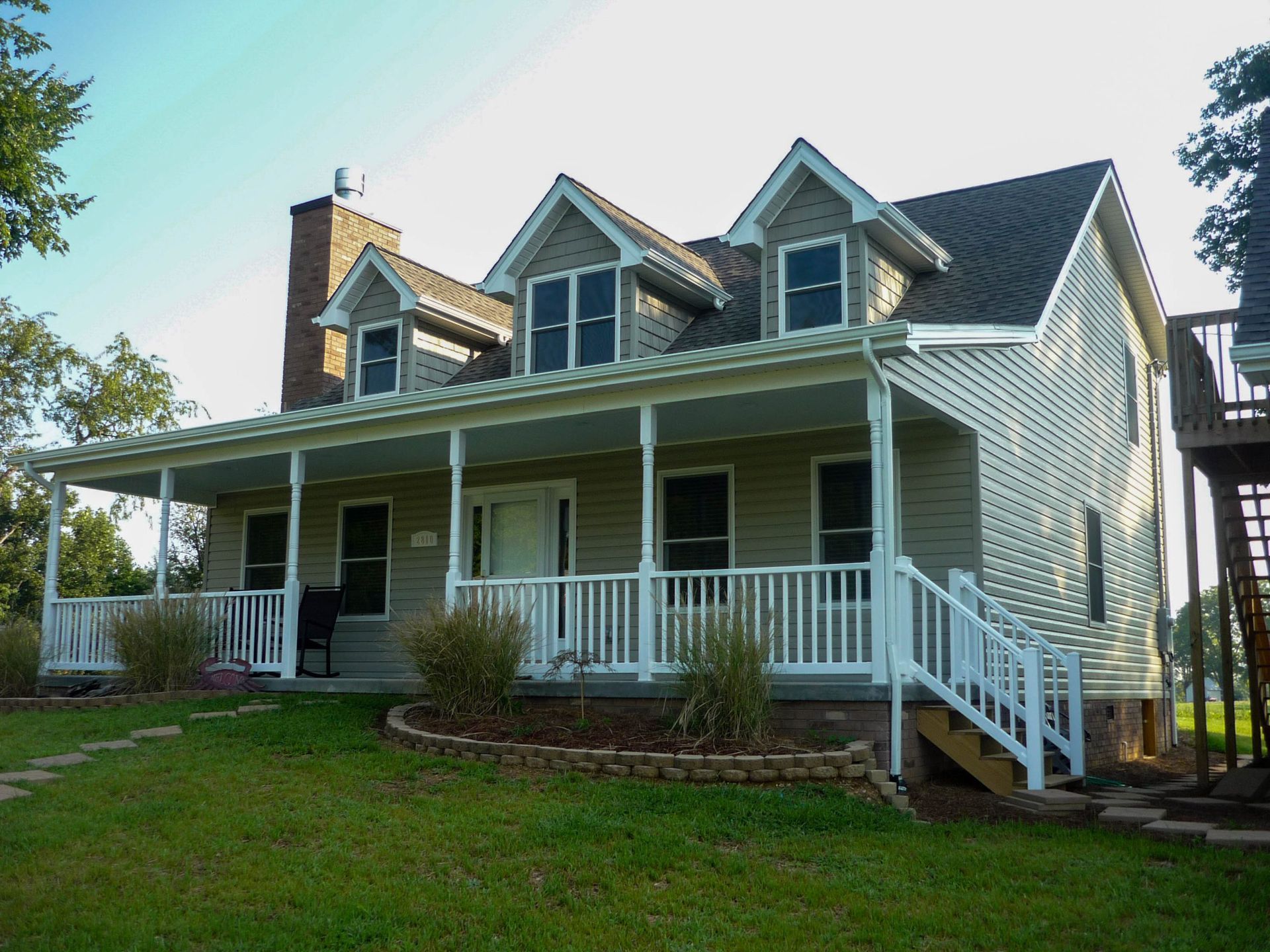 White two-story house with a front porch, white railings, dormer windows, and a green lawn