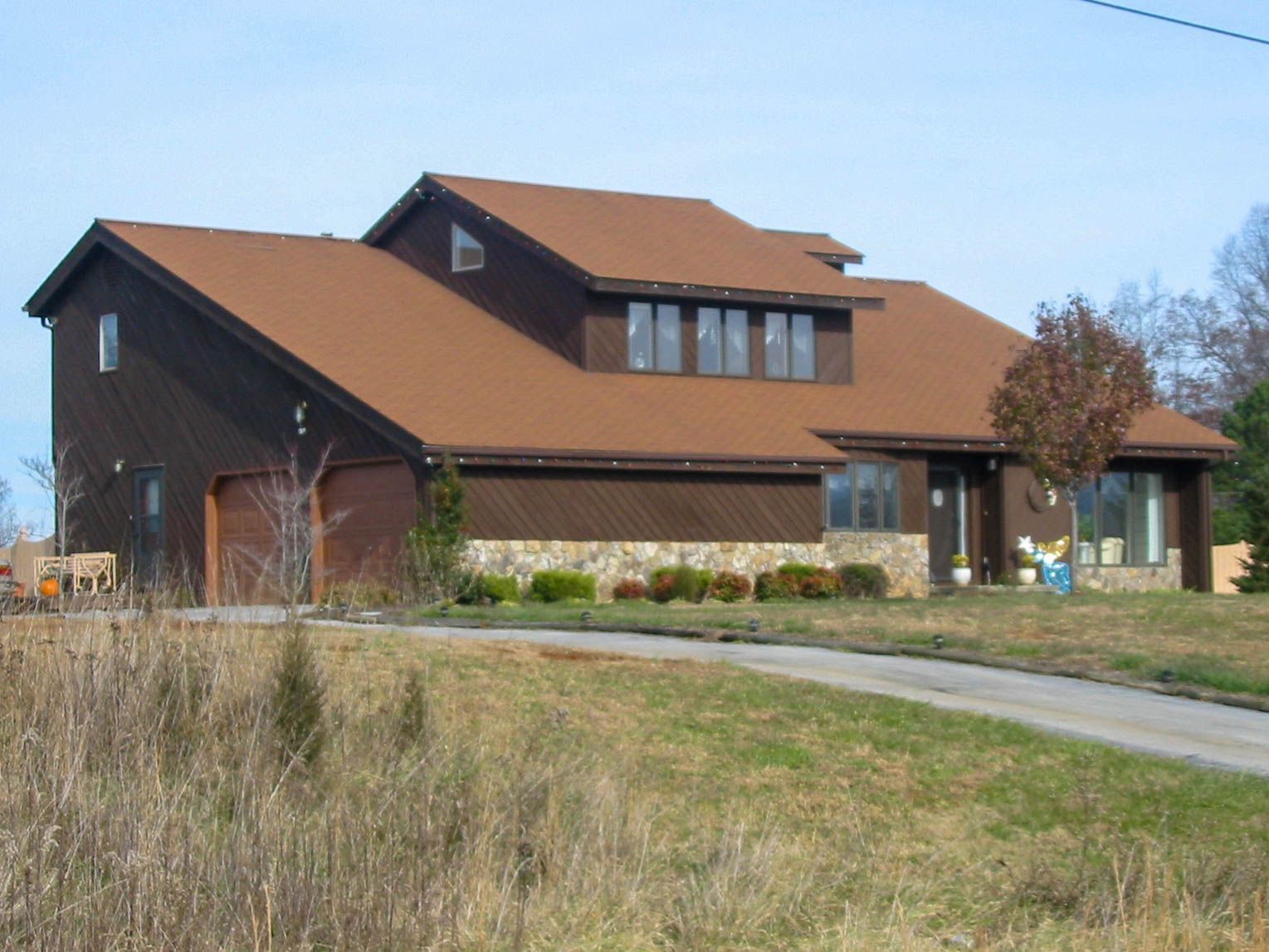 Brown house with a tan roof beside a grassy field and driveway under a clear sky