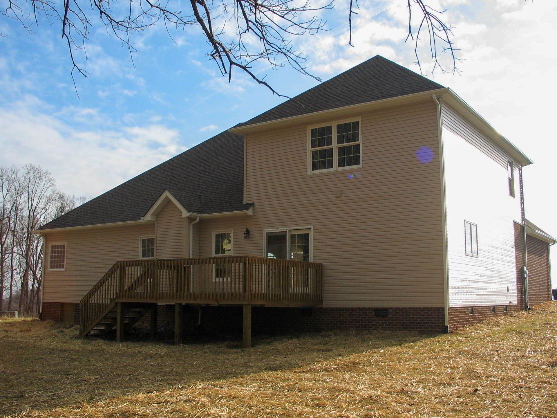 Side view of a tan house with a dark roof, wooden deck, and leaf-covered yard under a blue sky