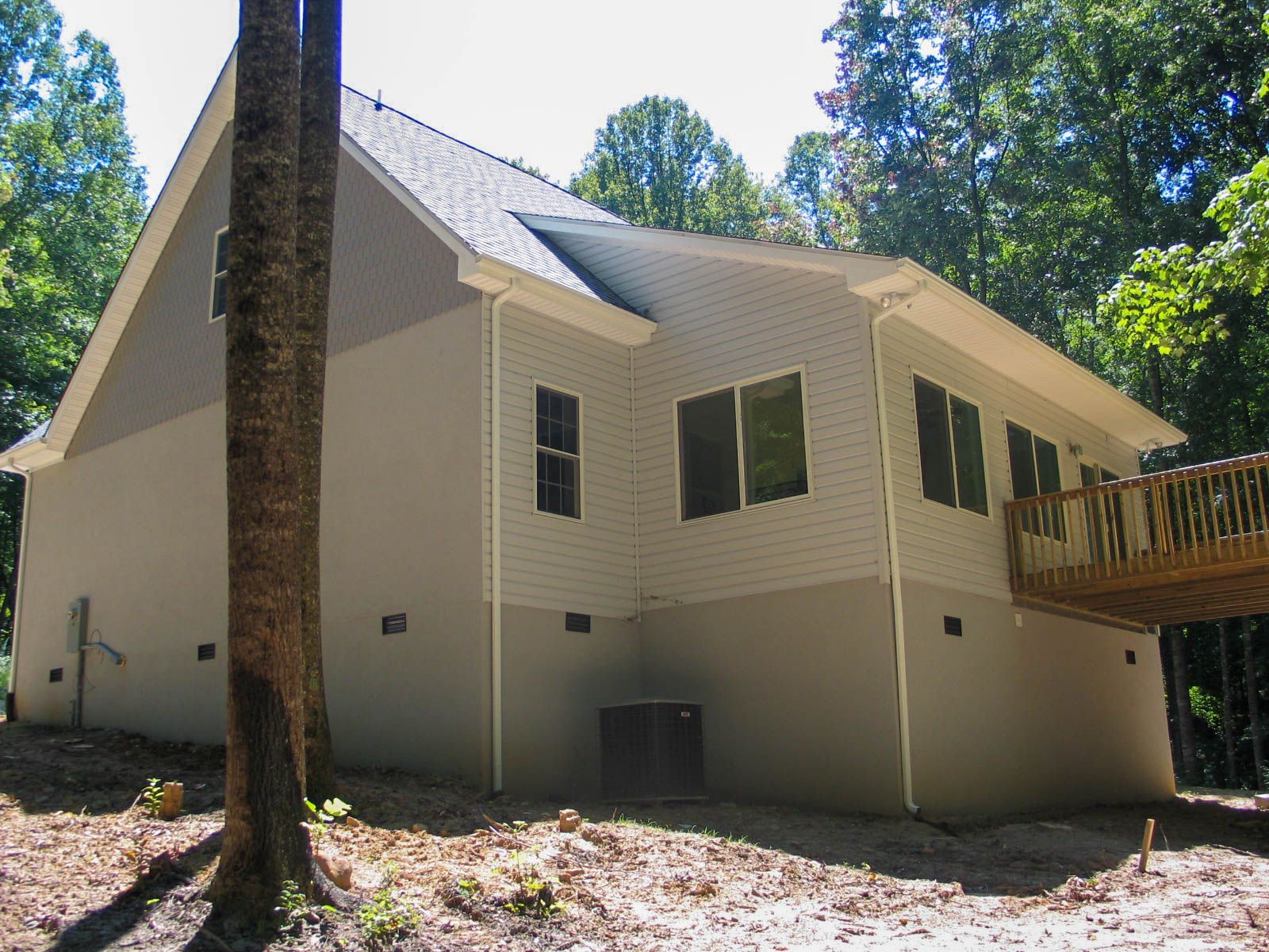 Small beige house with a steep roof, screened windows, and a wooden deck in a wooded yard