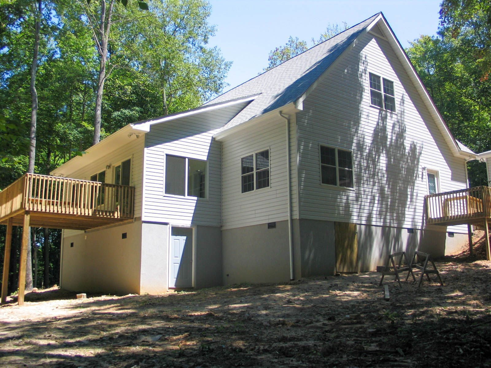 Wood-sided house with a steep roof and two wooden decks, surrounded by trees in a sunny wooded yard