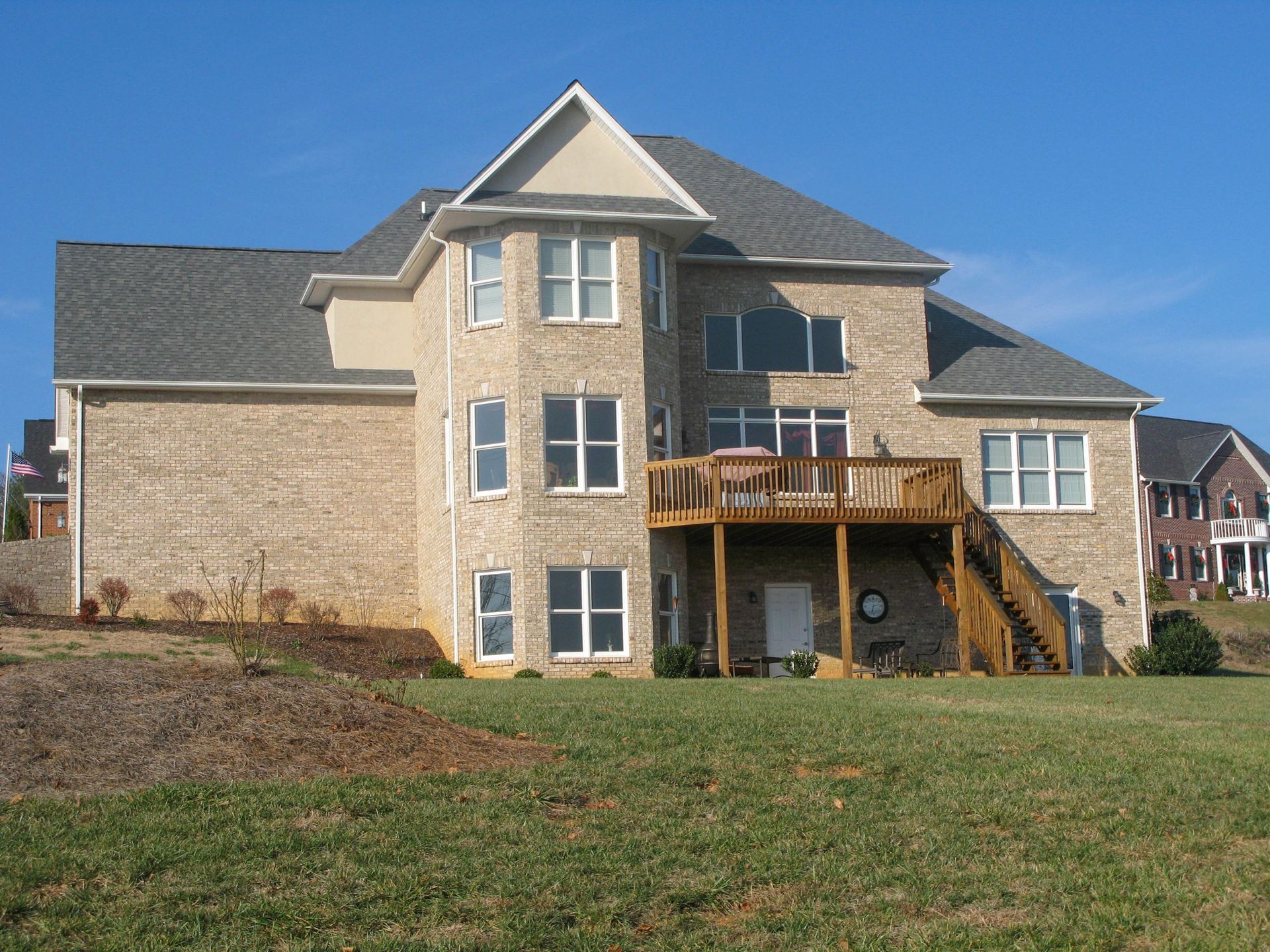 Two-story beige house with stone siding, wooden deck, and grassy yard under a blue sky