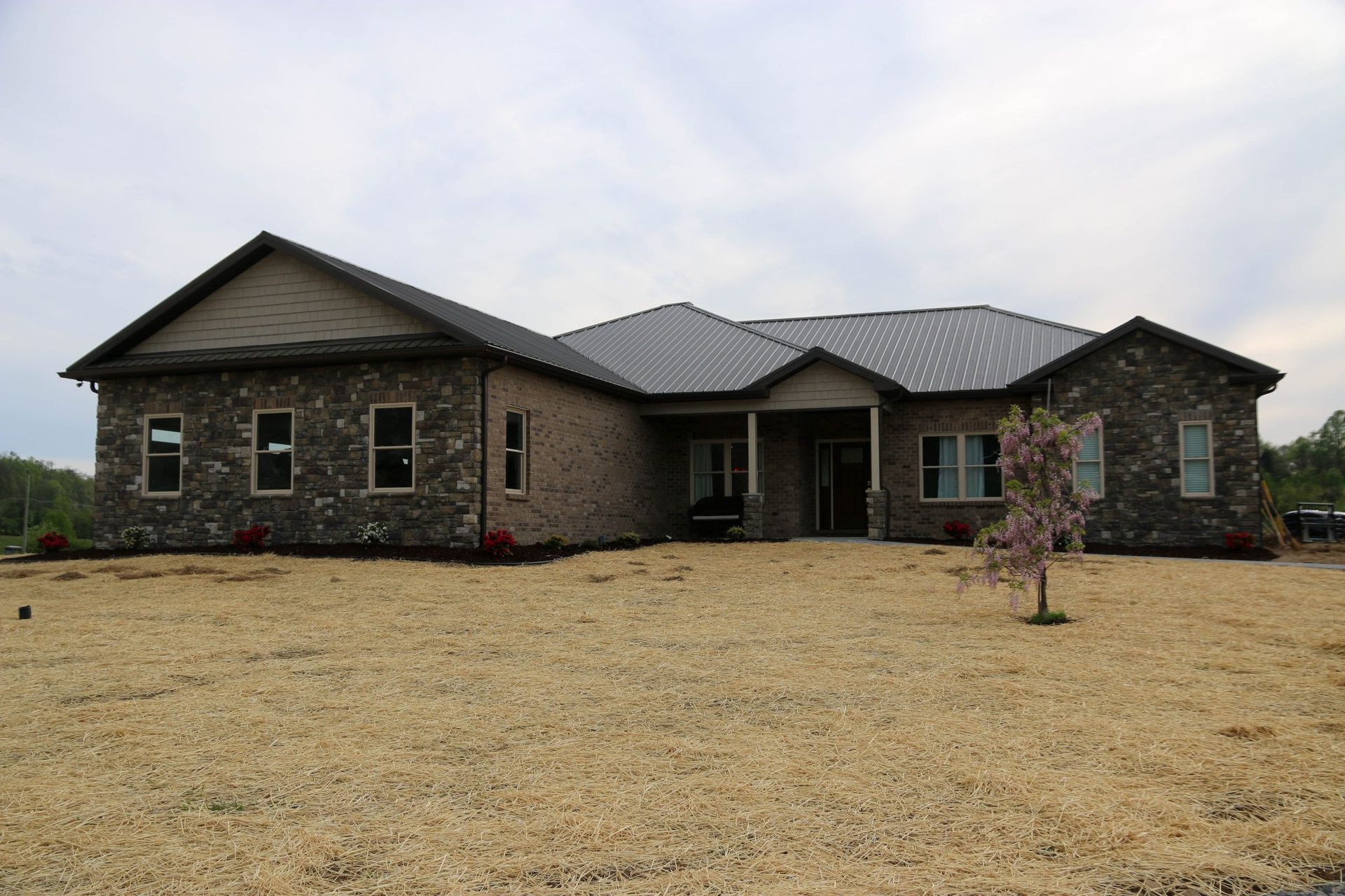 Single-story stone house with a dark roof on a dry lawn under an overcast sky