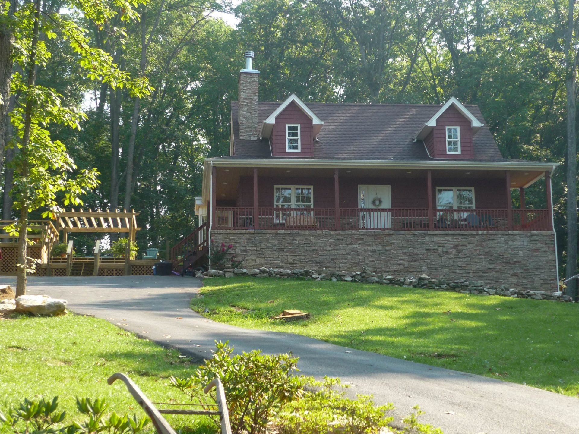 Small red cottage with stone porch in a wooded yard, with a curved driveway and green lawn