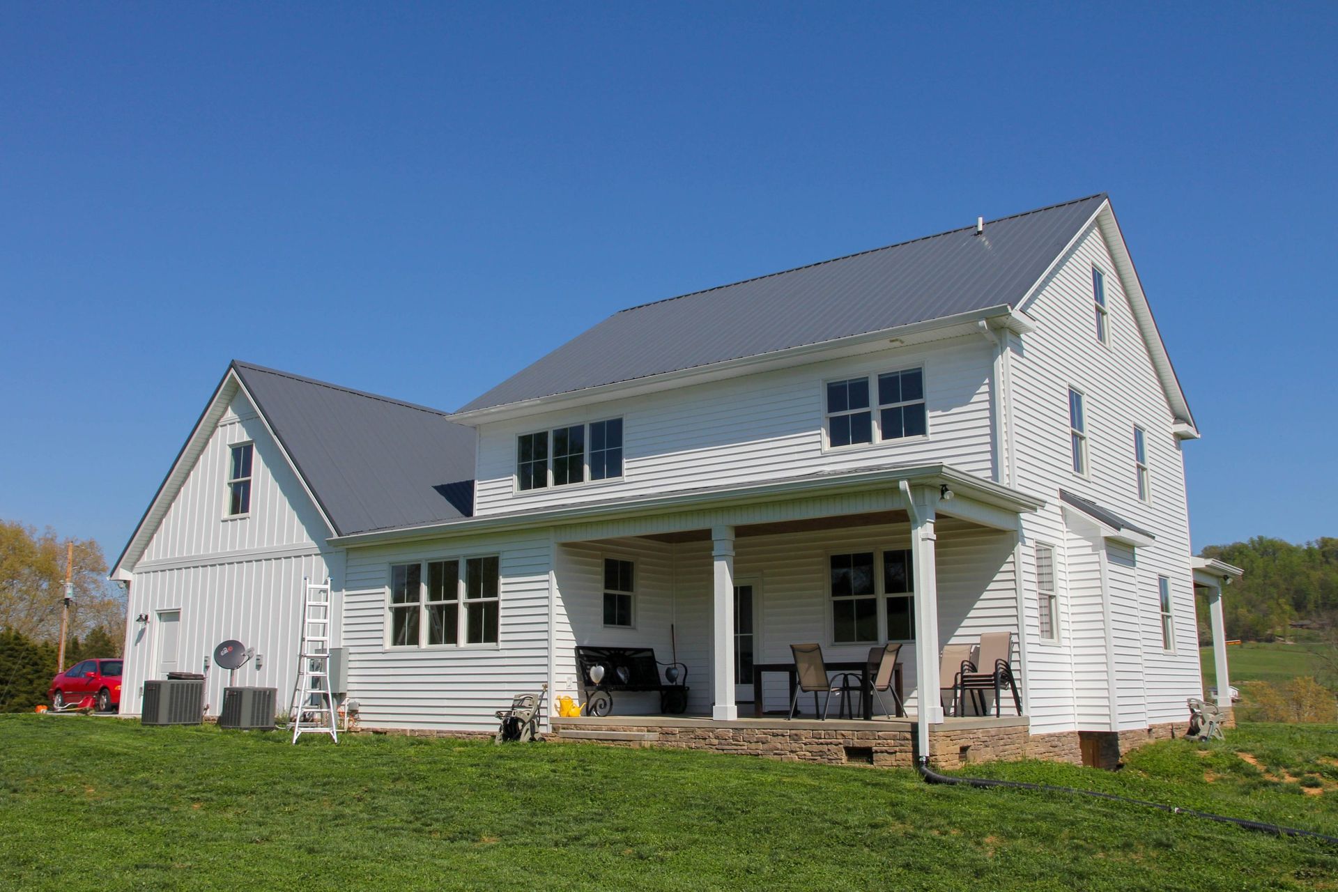 White farmhouse with gray roof and covered porch on a grassy hill under a clear blue sky