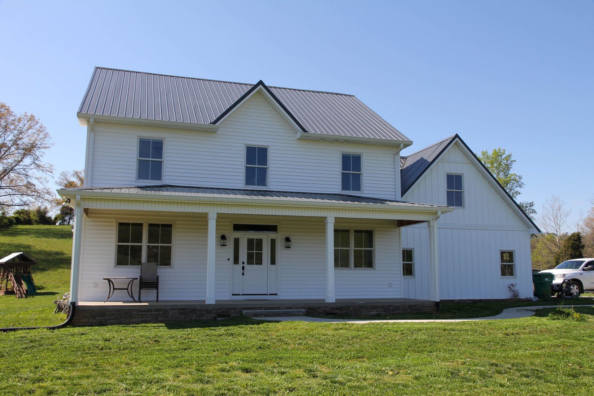 White farmhouse with porch and gray roof on a grassy lot under a blue sky