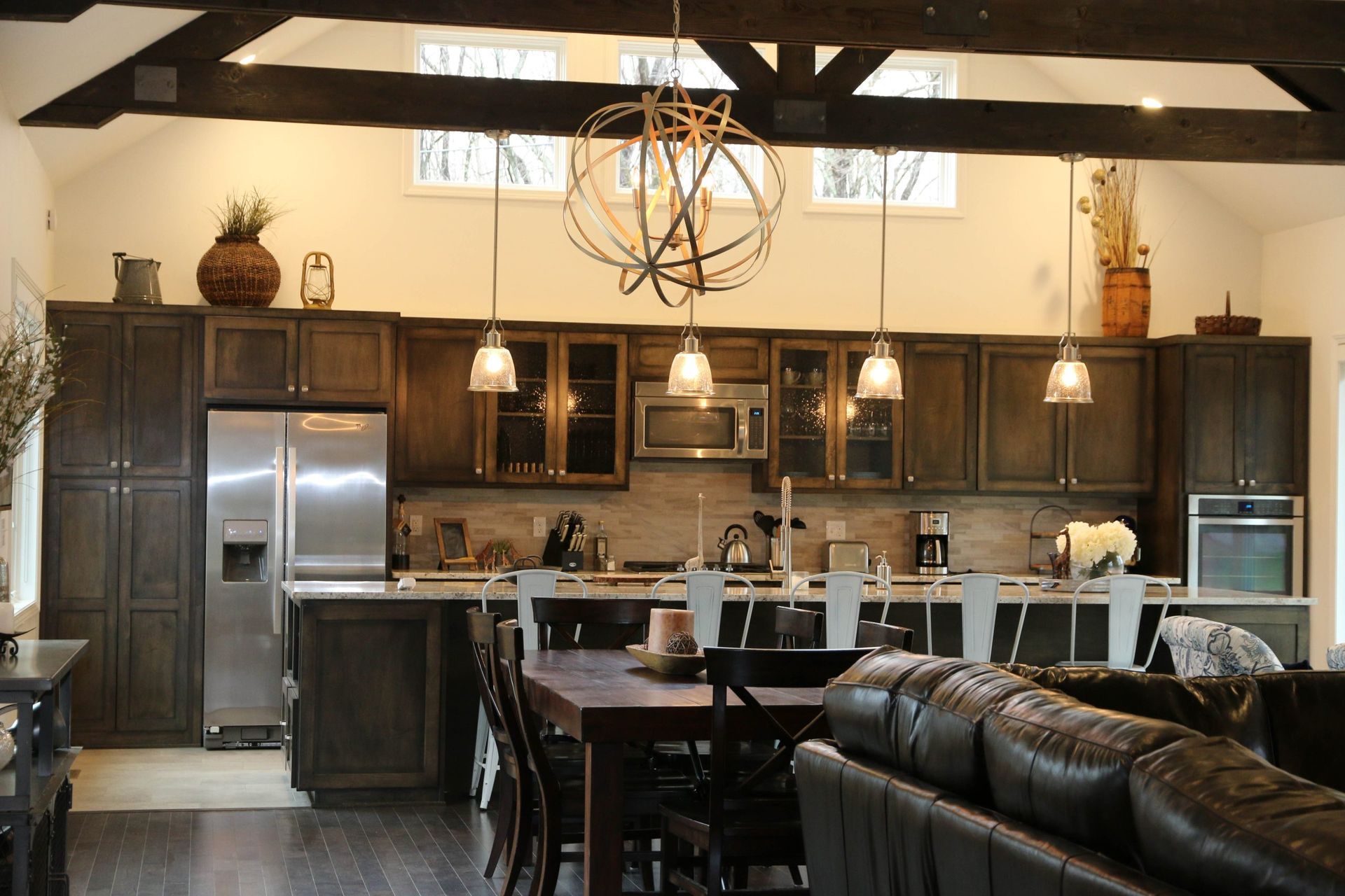 Modern open kitchen with dark wood cabinets, stainless appliances, high ceiling beams, and pendant lights.