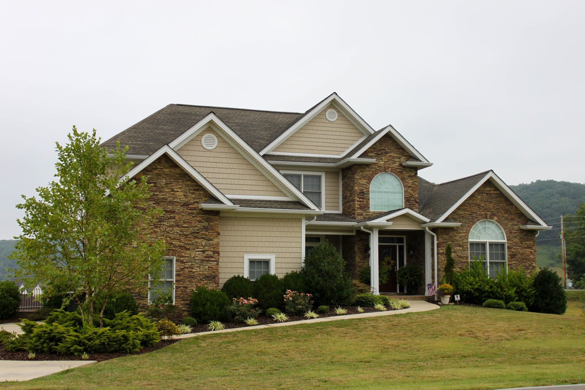 Large beige brick suburban house with a manicured lawn and landscaped garden under a cloudy sky