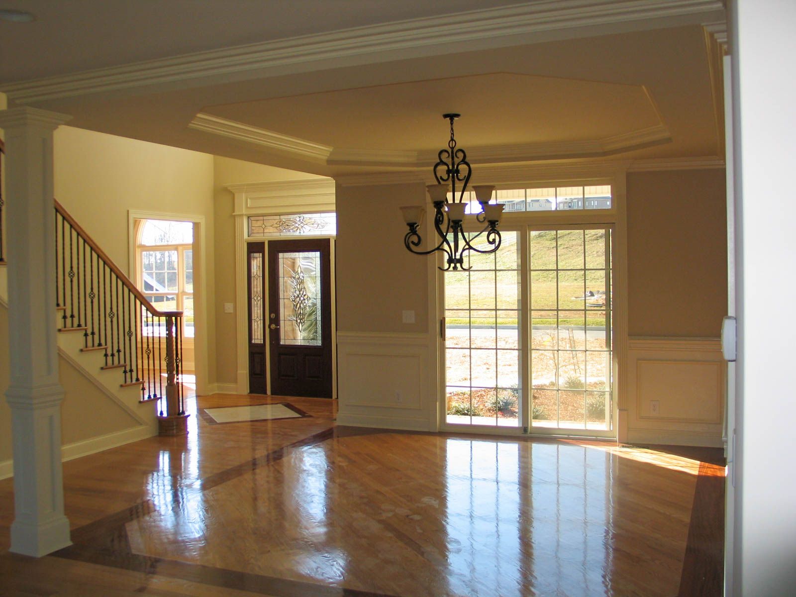 Empty sunlit living room with hardwood floors, chandelier, staircase, and large windows.