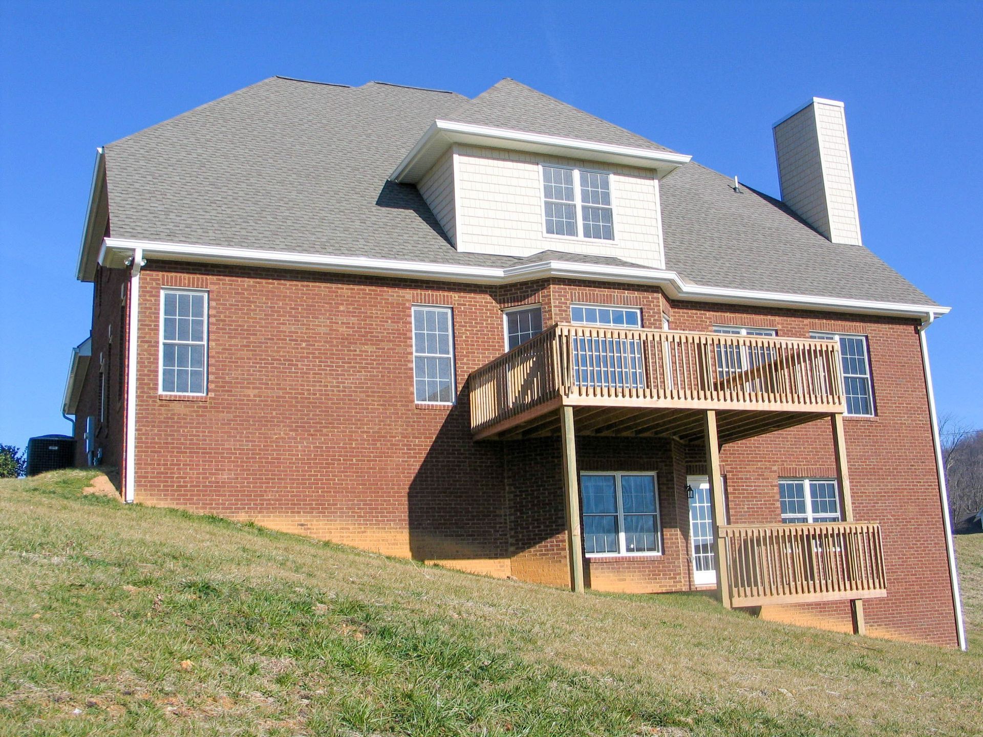 Brick house with a sloped roof, white trim, and wooden balconies on a grassy hill under a blue sky