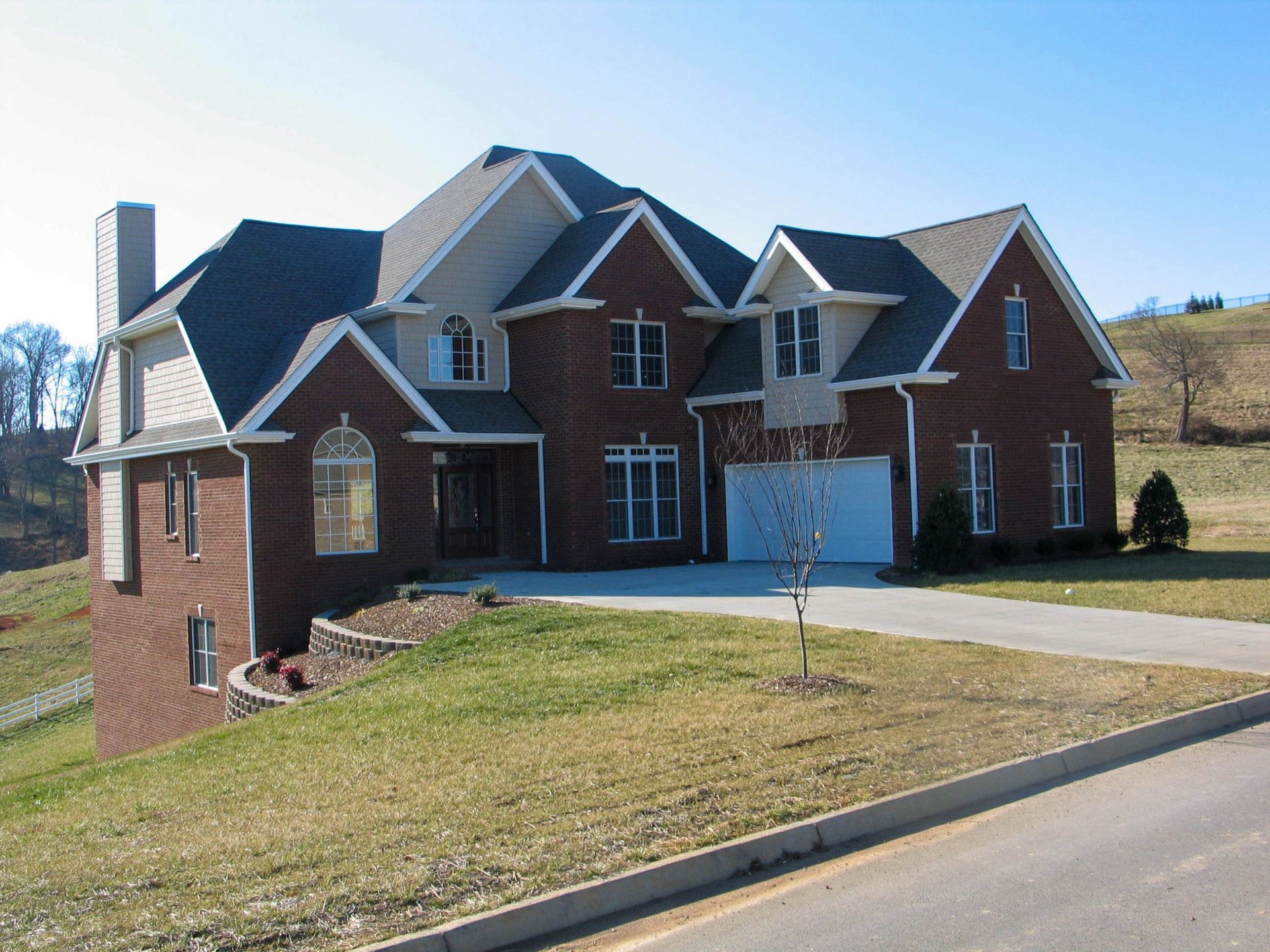 Large brick suburban house with a driveway, garage, and manicured lawn under a clear blue sky