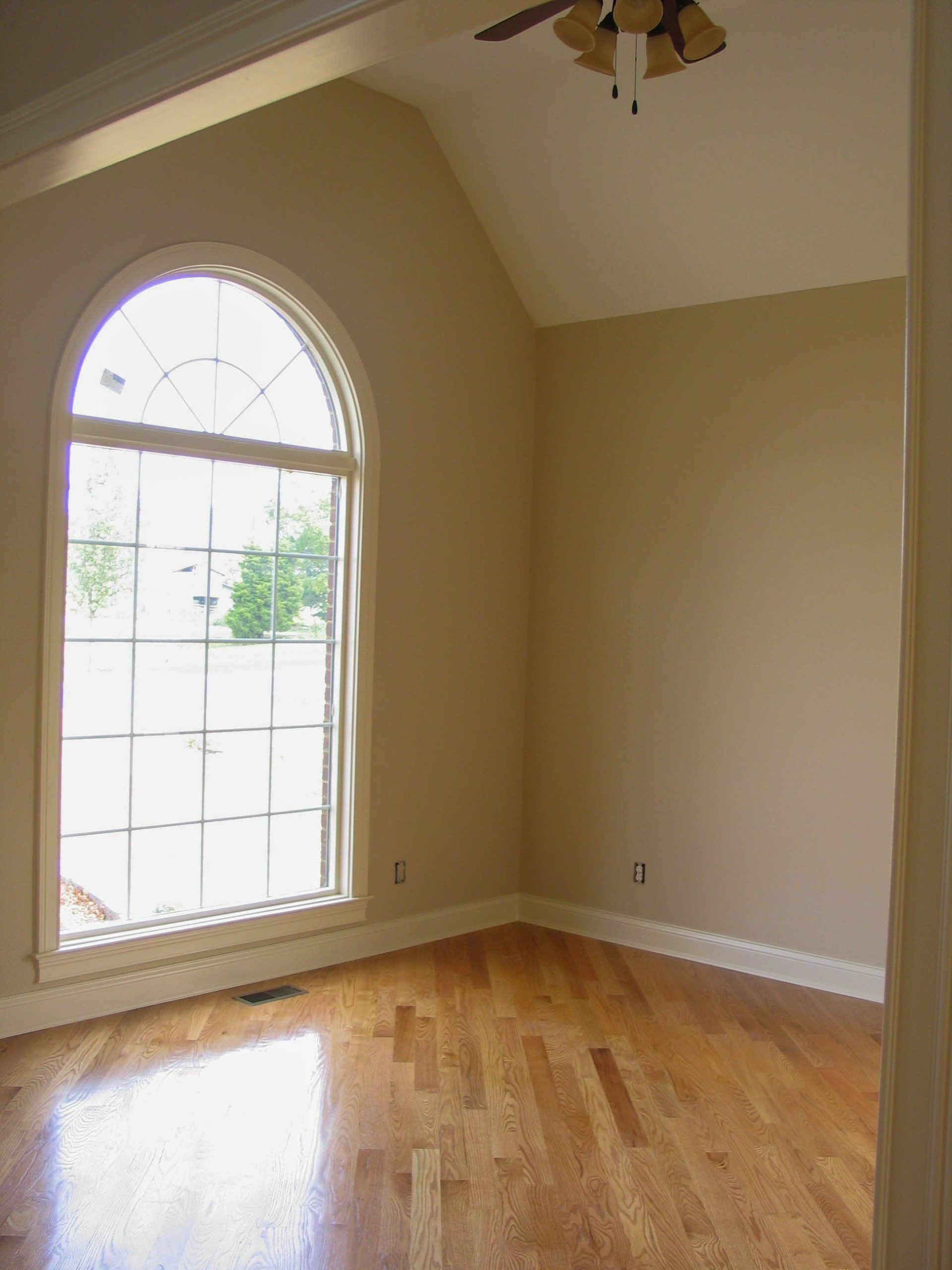 Empty beige room with hardwood floors and a large arched window letting in natural light
