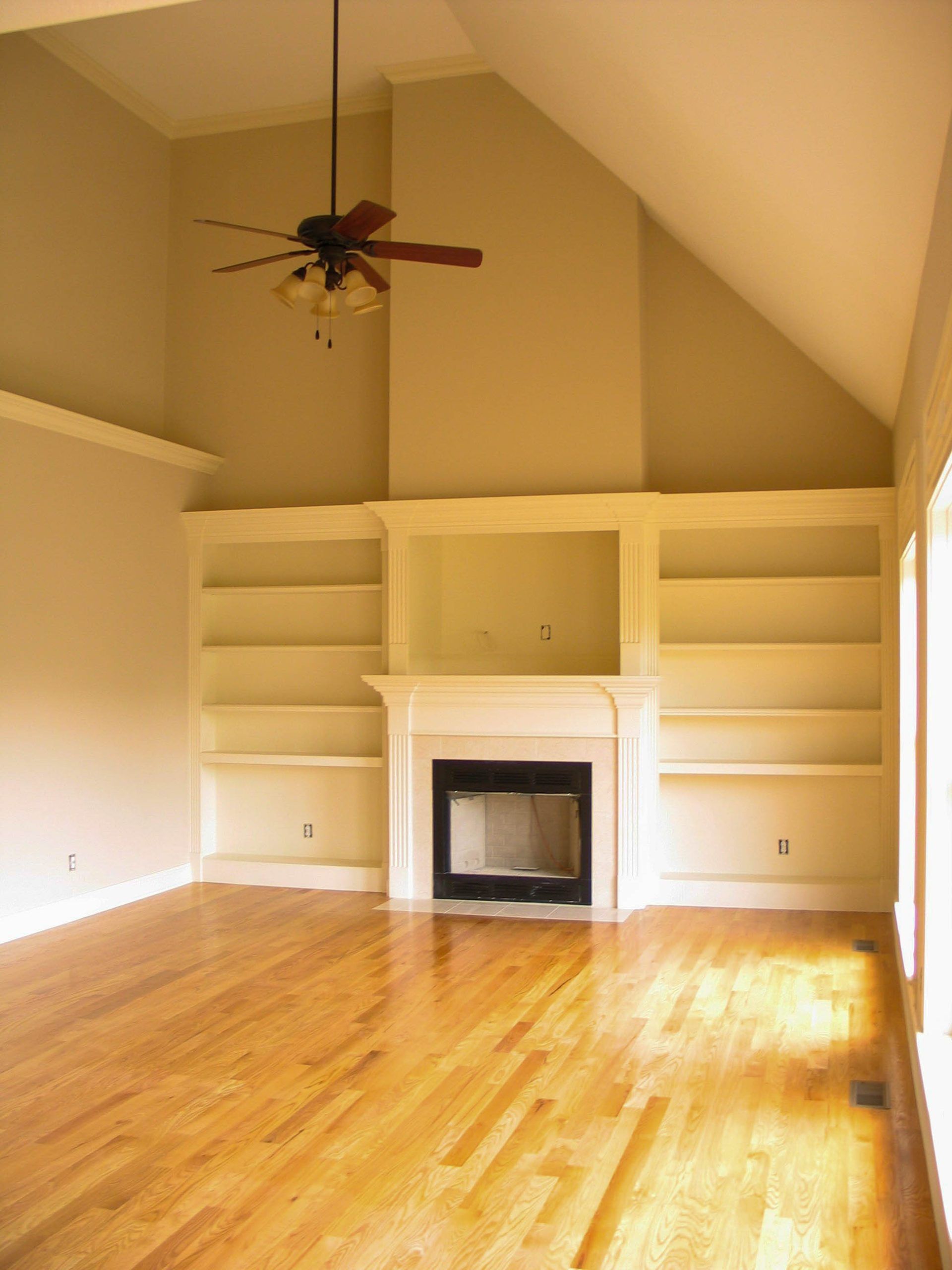 Empty living room with vaulted ceiling, hardwood floor, built-in shelves, fireplace, and ceiling fan