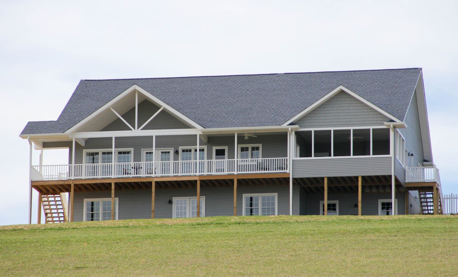 Large gray house with a covered porch and balcony on a grassy hill under a cloudy sky
