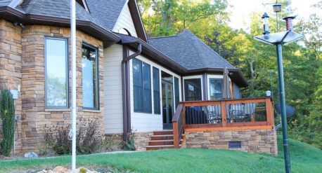 Stone house exterior with large windows, wooden deck, and black lamp posts on a grassy slope