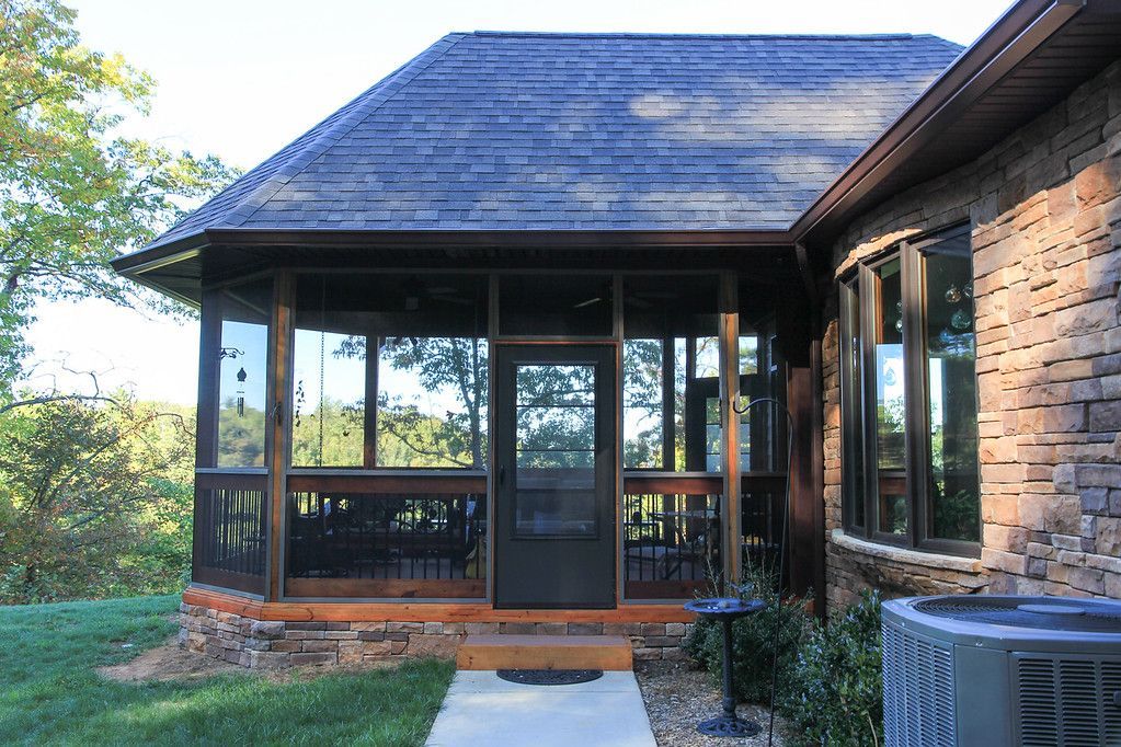 Front porch with screened windows, dark roof, brick wall, and air conditioner unit beside a grassy yard.