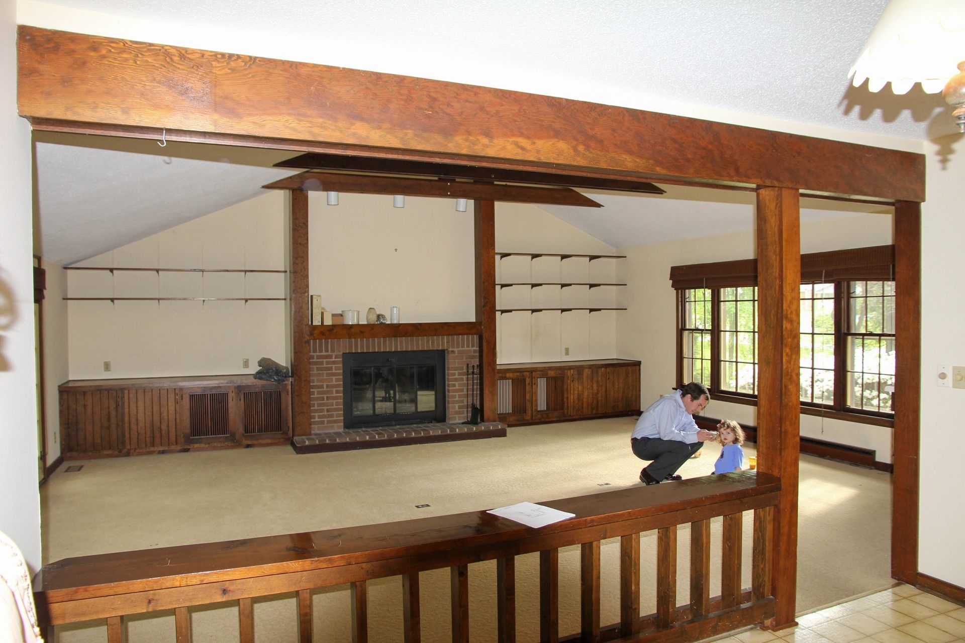 Open living room with fireplace and wood beams; person sitting on a bench near large windows.
