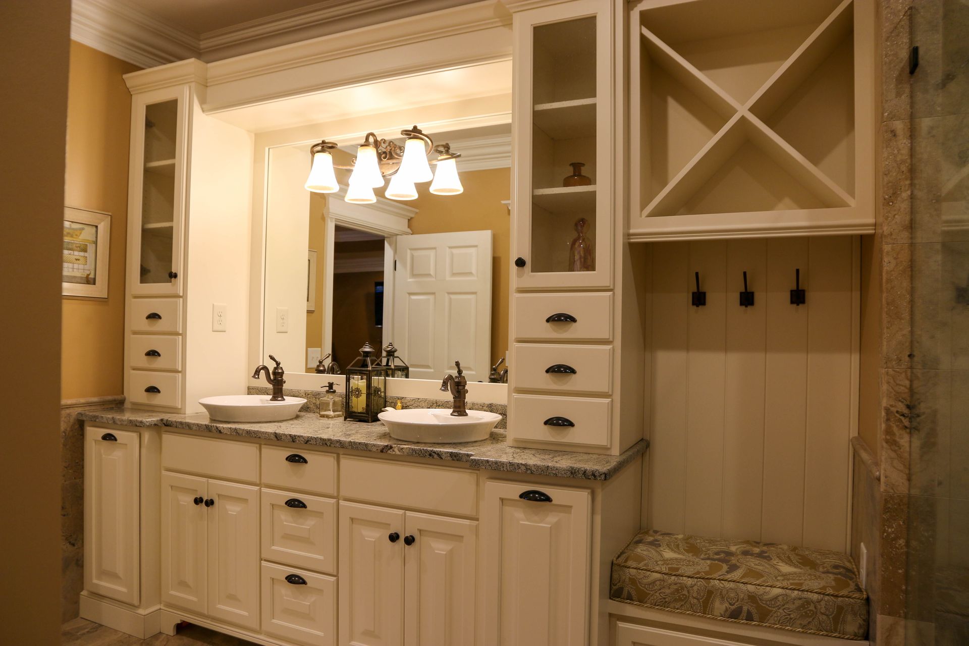 A bright, off-white bathroom vanity with dual sinks, overhead lighting, tall cabinets, and a cushioned seating nook.