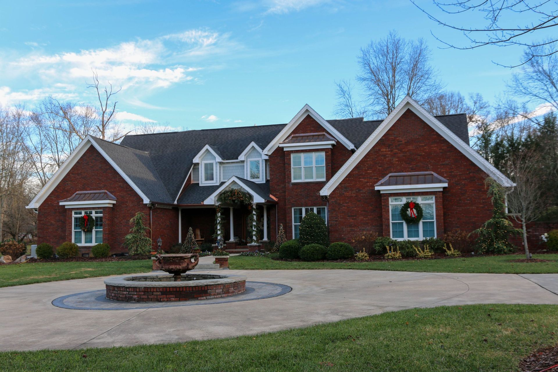 Large brick suburban house with a circular driveway, front porch, and landscaped yard under a blue sky