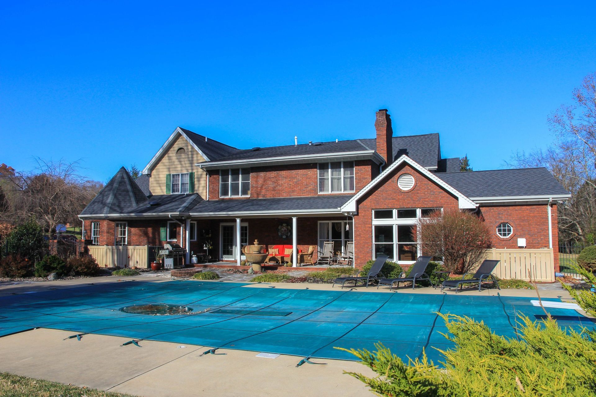 Brick house with red roof and covered porch beside a blue swimming pool under a clear sky