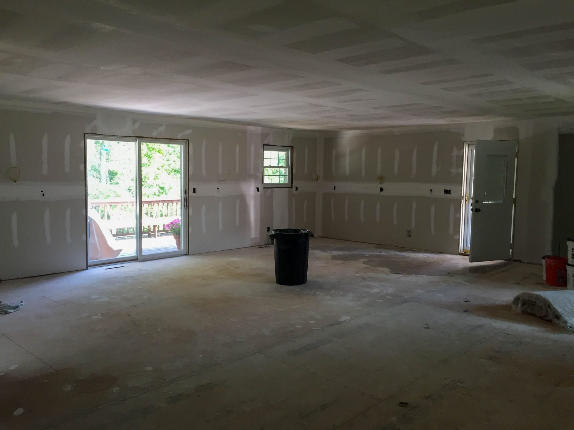 Empty unfinished room with drywall, exposed floor, and sunlight through open doors and windows.