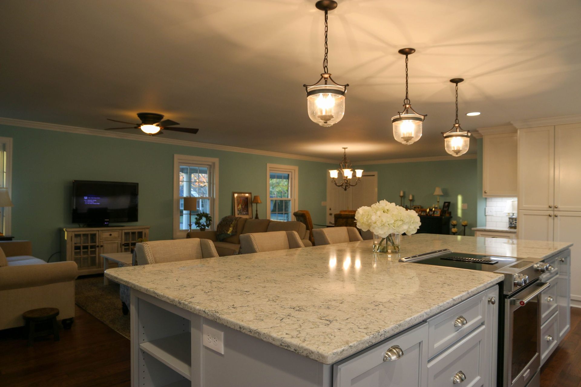 Bright modern kitchen with large island, pendant lights, and open view into a living room with a fireplace and TV.