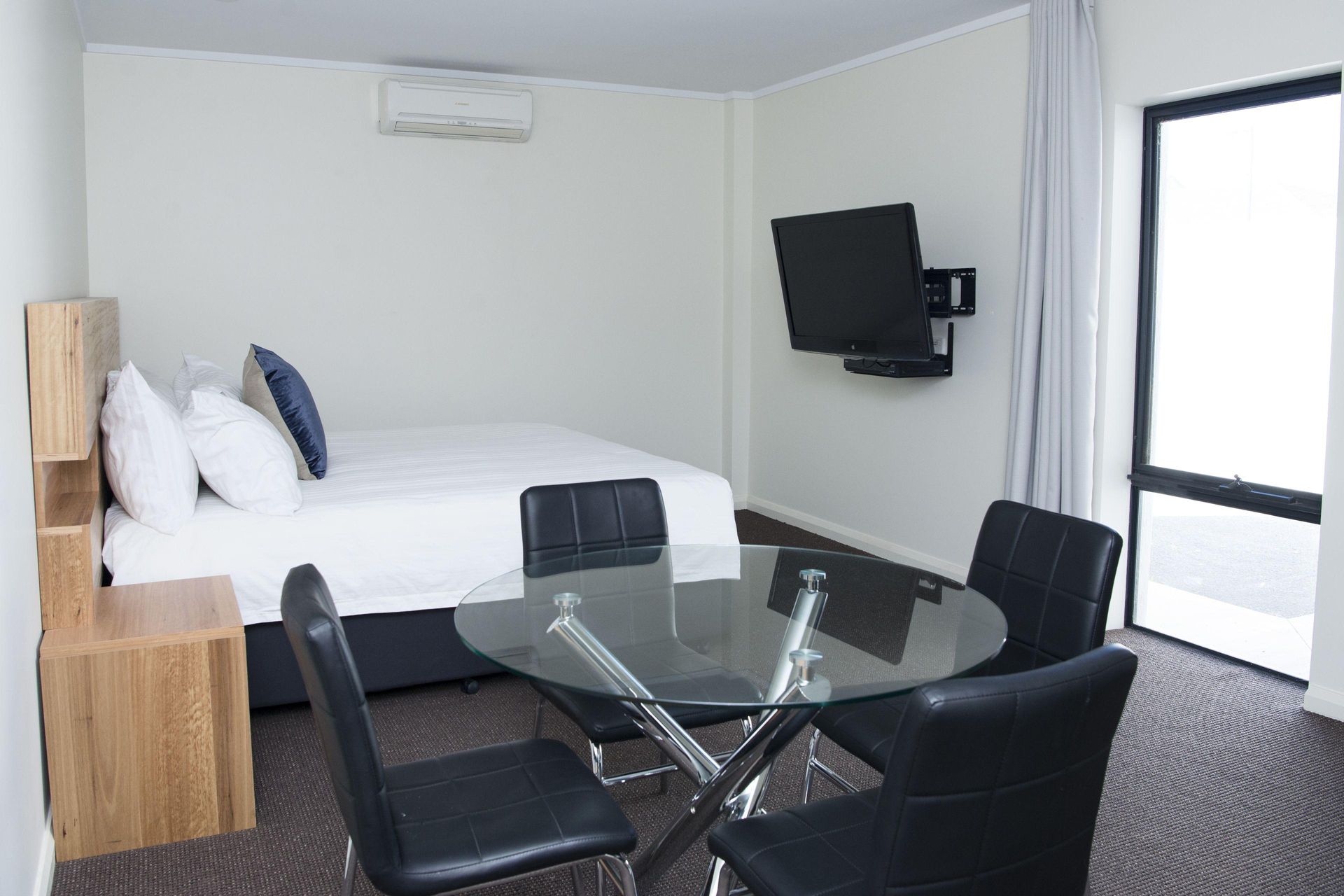 Modern bedroom with white bed, glass table and black chairs, wall-mounted TV, and large window.
