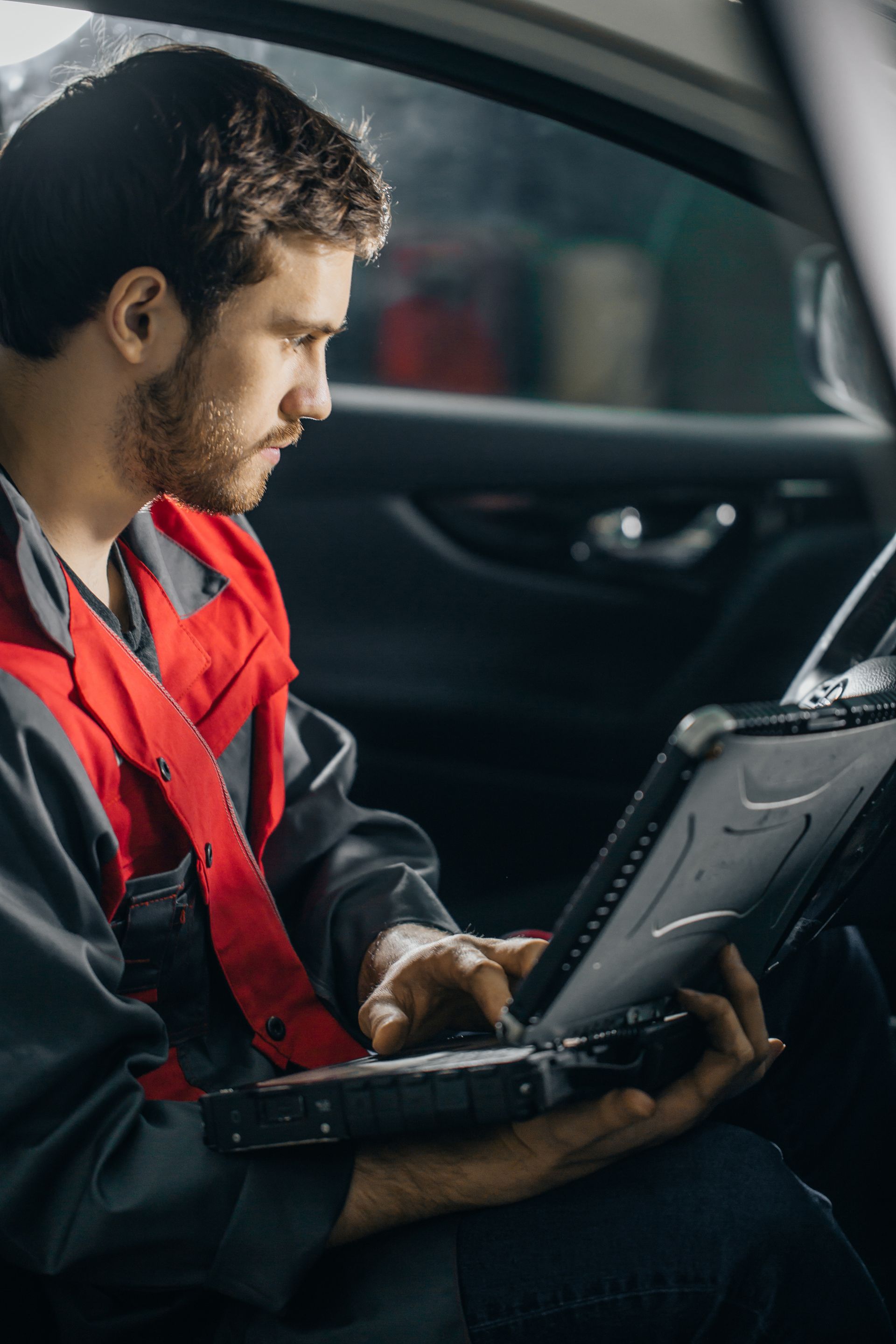 Mechanic in red jumpsuit using a laptop inside a car, likely diagnosing a vehicle issue | Calgary Auto and Alignment