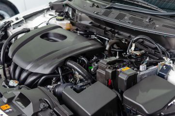 Car engine bay with black plastic cover and visible battery and hoses under the hood