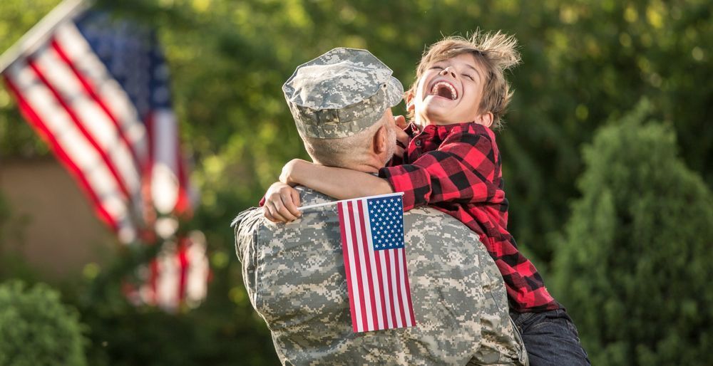 Soldier in camouflage uniform hugs laughing child outdoors, American flags in background.