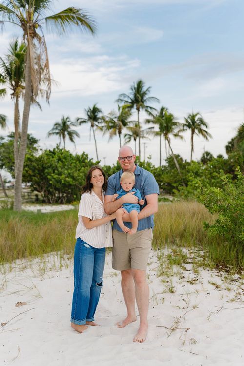 Family standing on a sandy beach with palm trees, holding a baby under a bright sky.