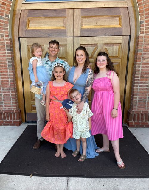 A family of six stands smiling in front of large wooden doors, dressed in colorful clothing for a festive occasion.