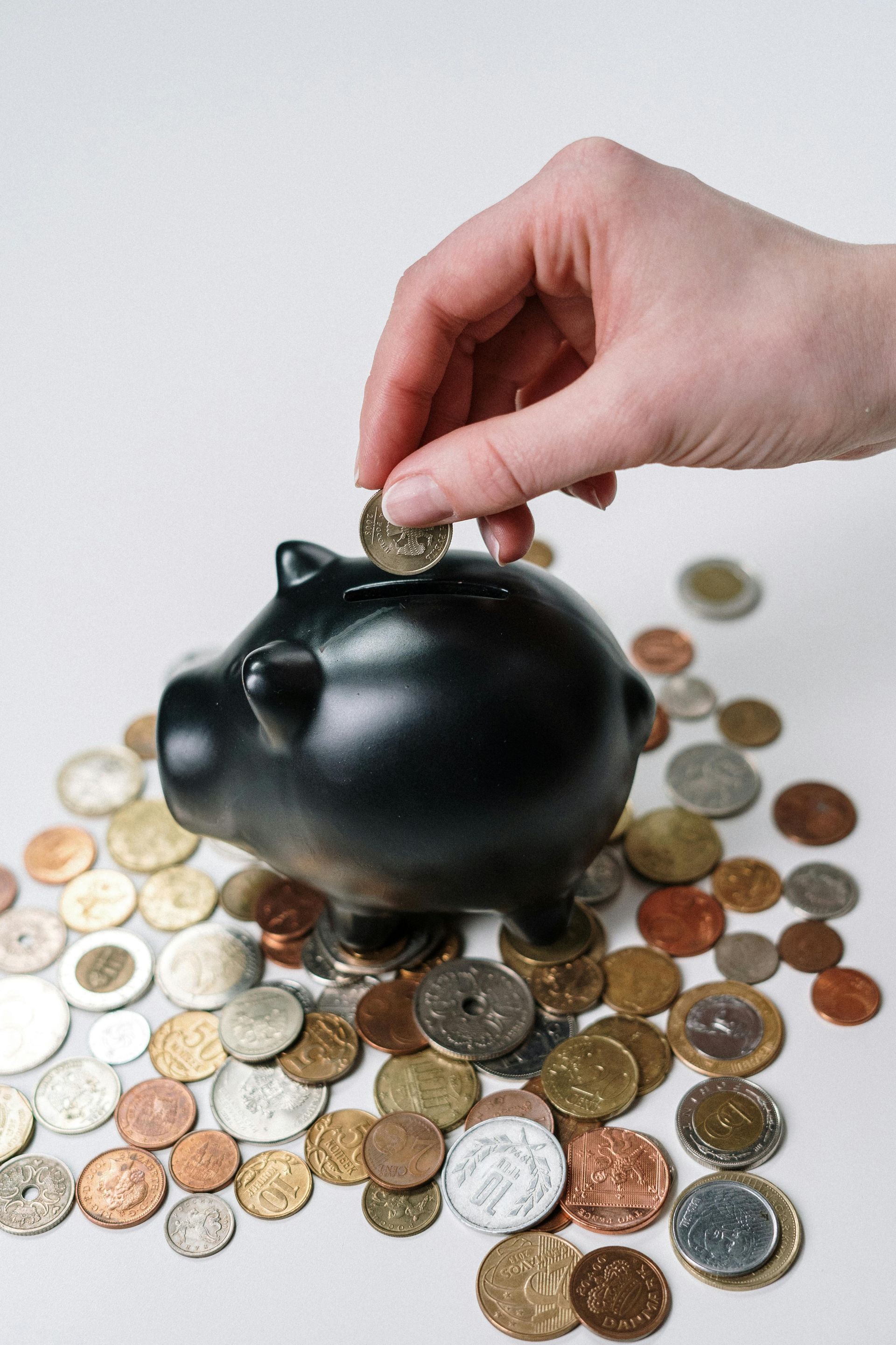 Hand placing a coin into a black piggy bank surrounded by various coins.