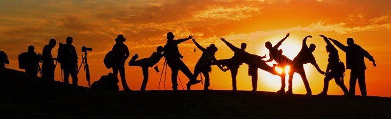 A group of people are standing in front of a sunset holding hands.