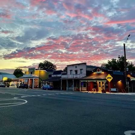 Small town street at dusk with lit storefronts and a colorful pink-blue cloudy sky.