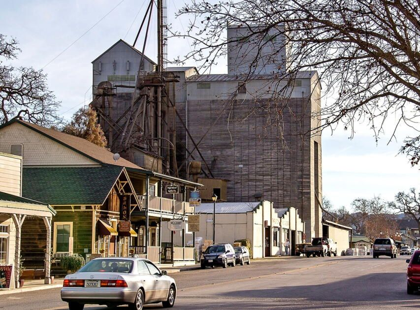 Small-town street with parked cars, storefronts, and a large industrial building wrapped in scaffolding